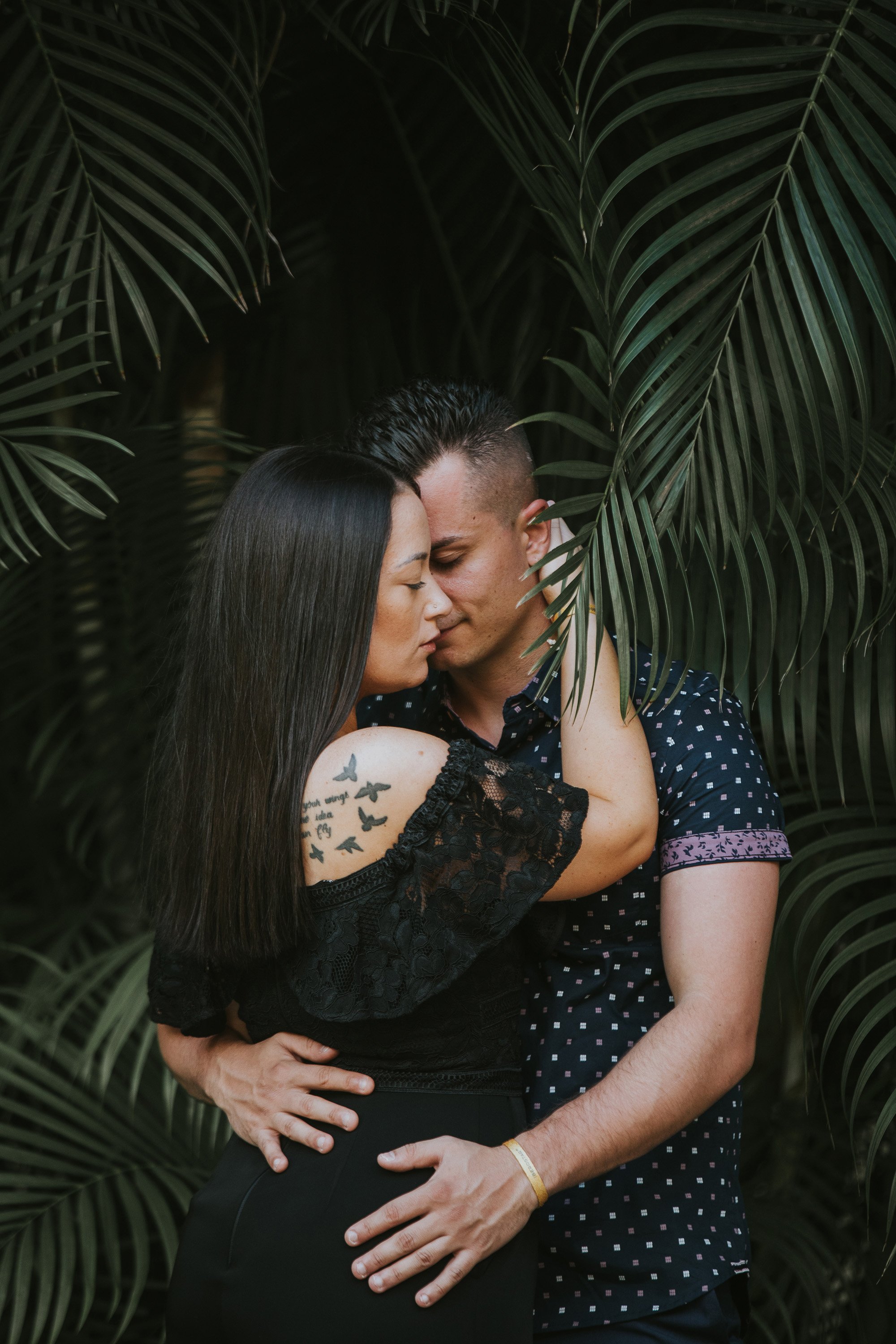 A couple shares an intimate embrace in a lush tropical forest, with the woman's tattoo visible on her shoulder, symbolizing their deep connection and love.