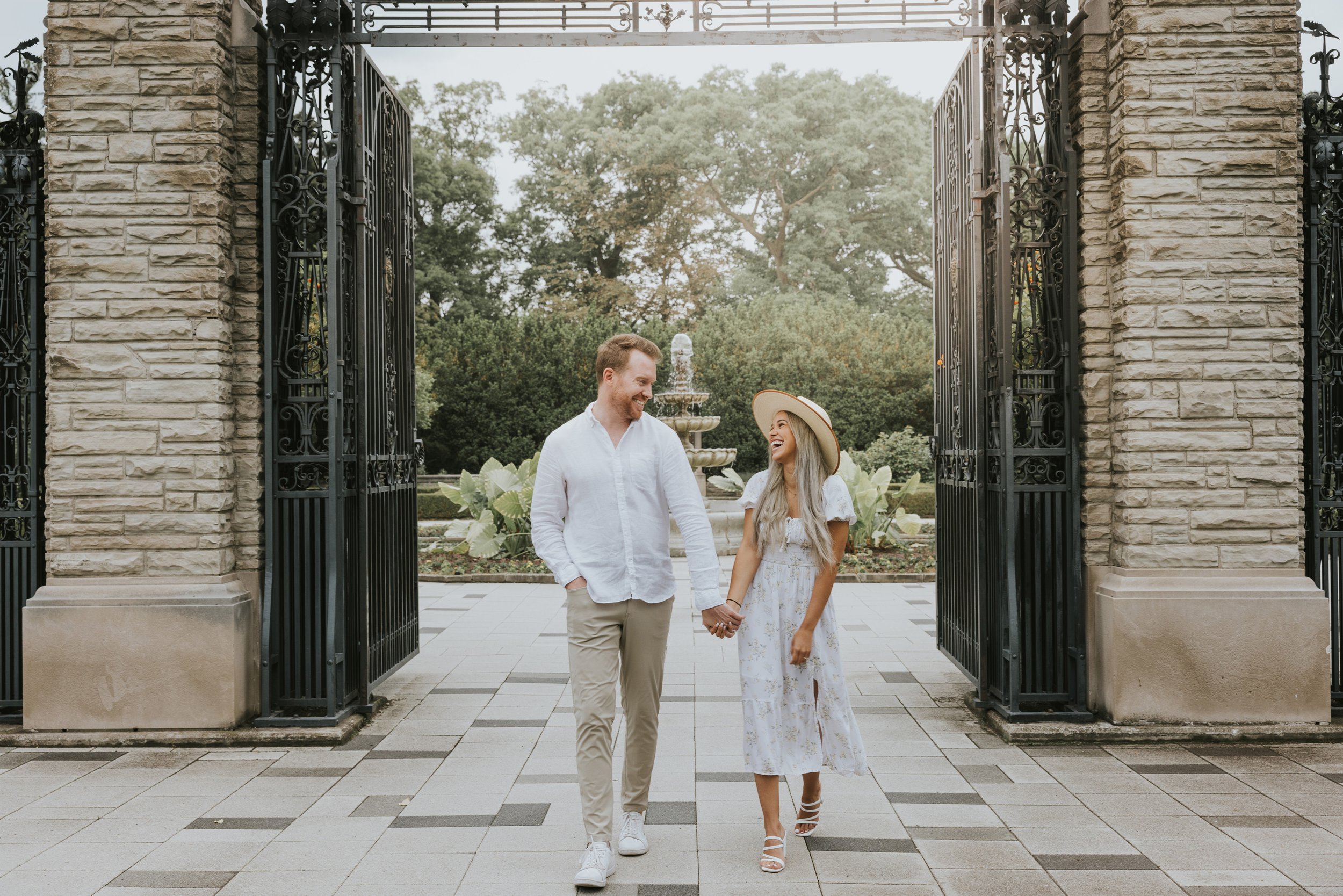 A couple walks hand in hand through the grand gates of a garden, sharing a joyful moment as they enter the beautiful outdoor space.
