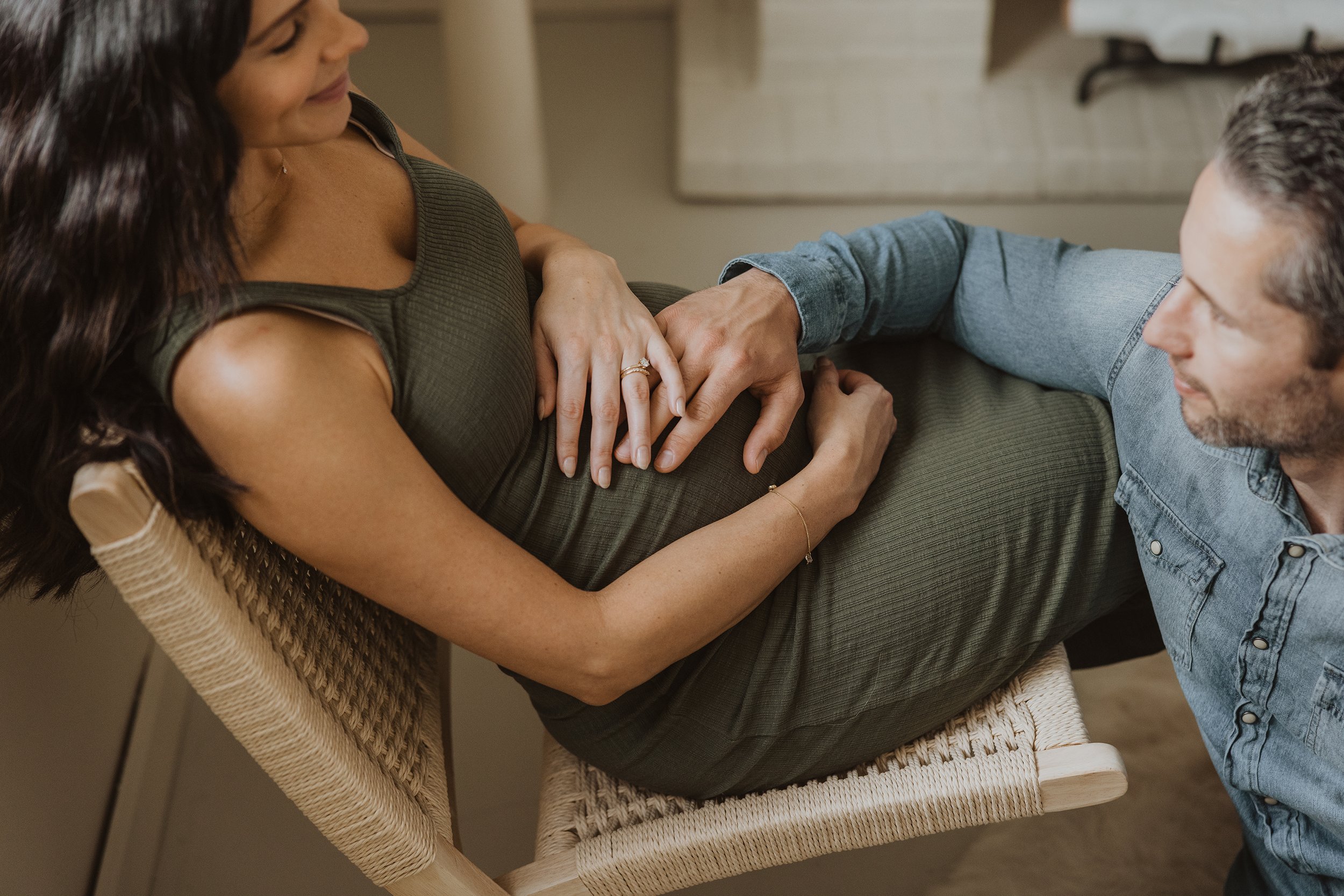A tender moment captured between a husband and his pregnant wife, with their hands resting on her baby bump as she sits in a chair."