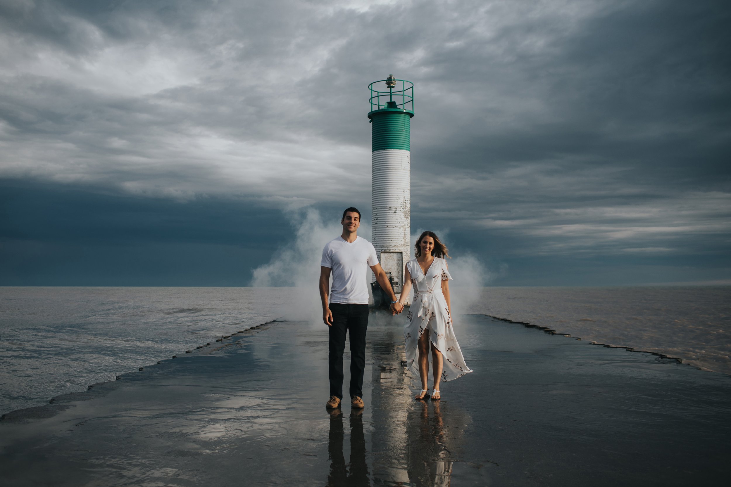 Couple walking hand-in-hand on a pier beside a lighthouse, with dramatic clouds and mist adding to the romantic atmosphere.