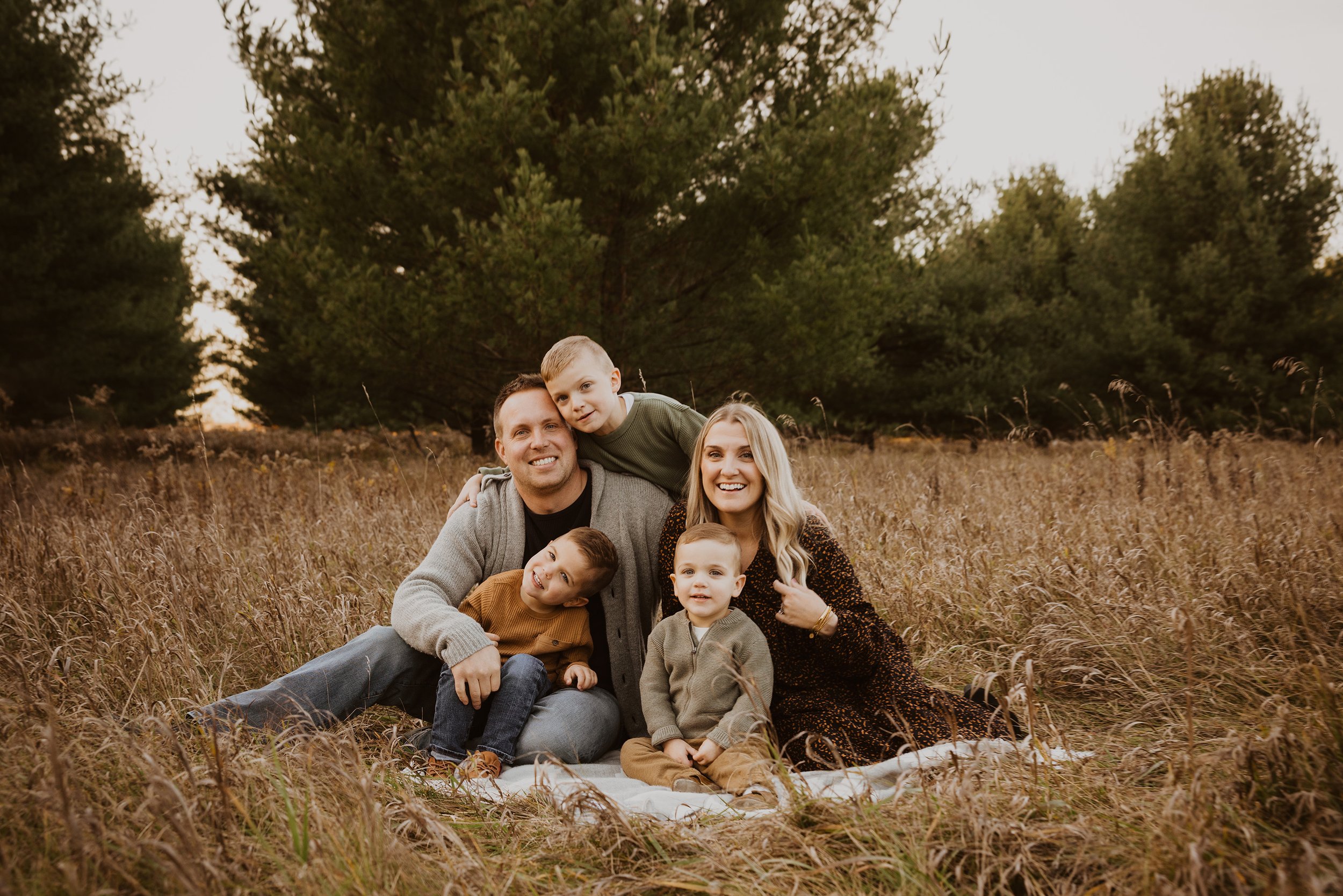 A family of five sitting close together in a field, surrounded by tall grass and trees, sharing a warm, candid moment during a nature photography session.