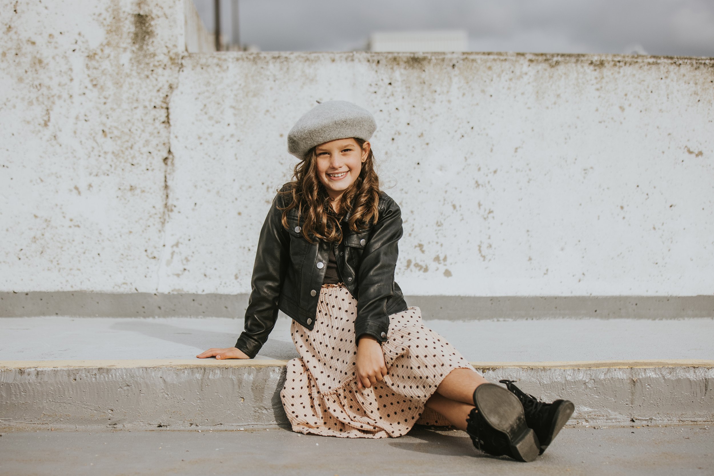 A young girl sitting on a concrete surface, dressed in a stylish outfit with a leather jacket and beret, smiling confidently at the camera.