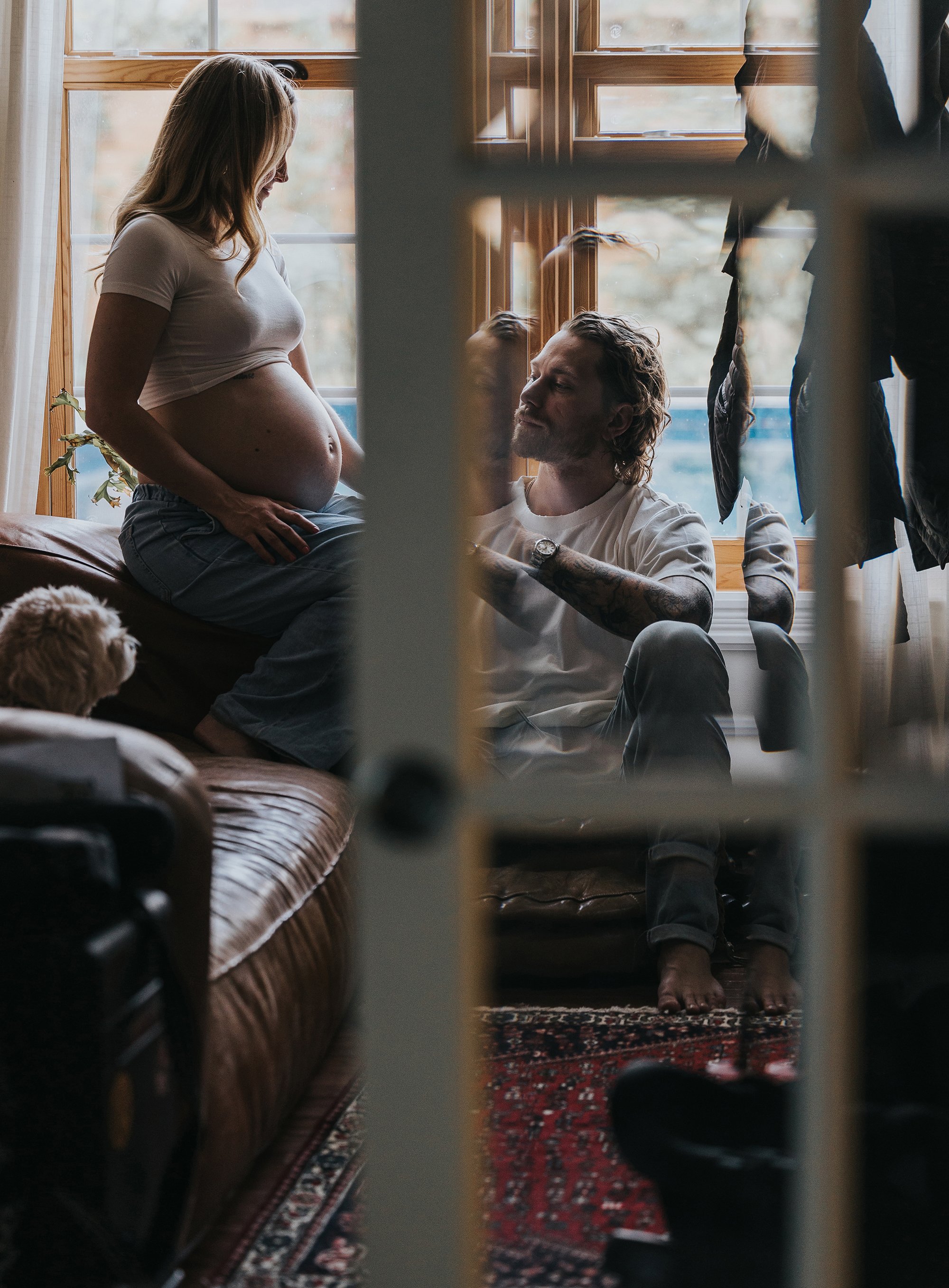 An intimate moment of a pregnant woman sitting on a windowsill, with her partner kneeling beside her, reflected through a nearby glass door.