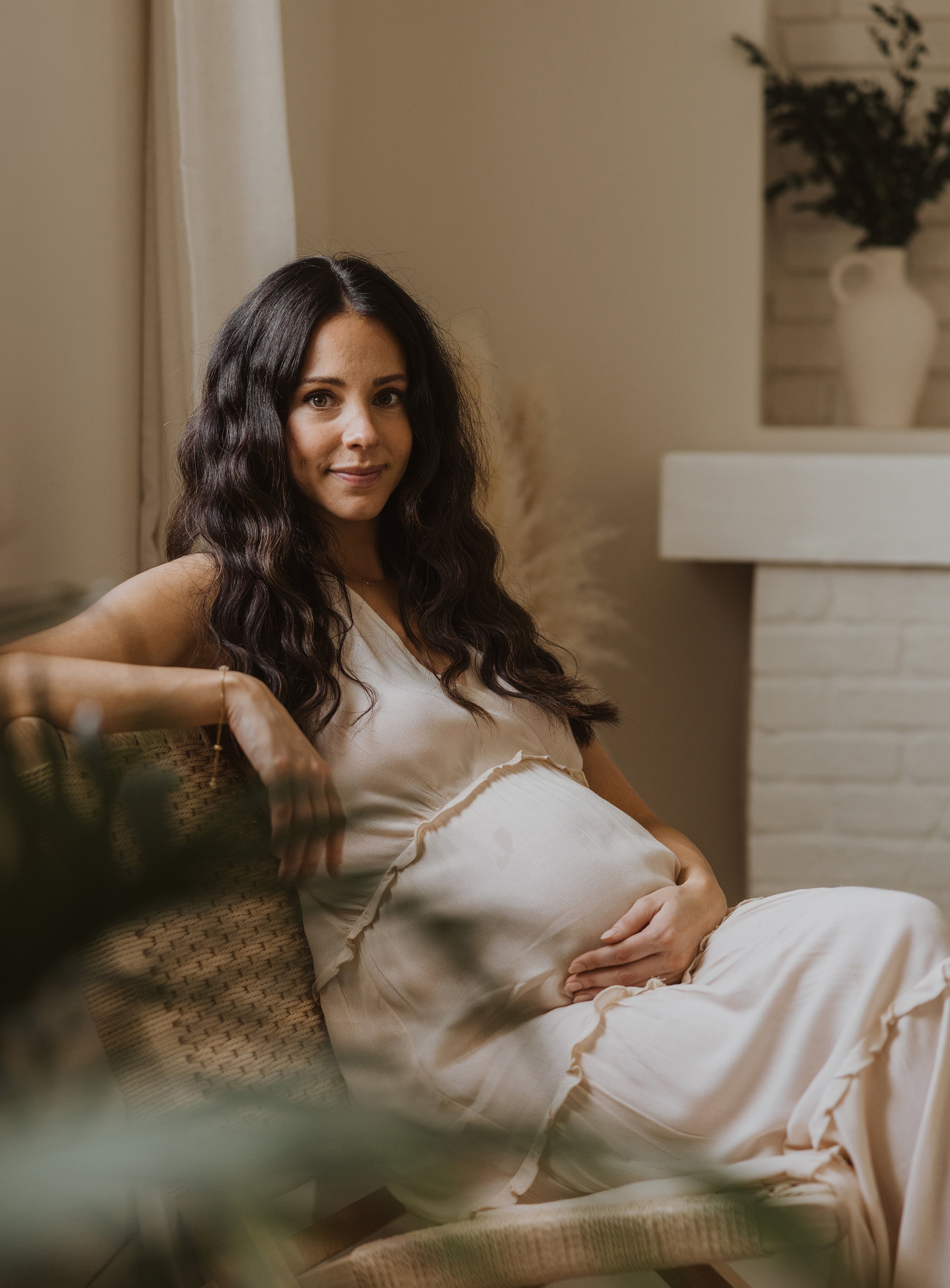 Pregnant woman sitting gracefully in a wicker chair, cradling her belly, with a serene expression in a naturally lit room.