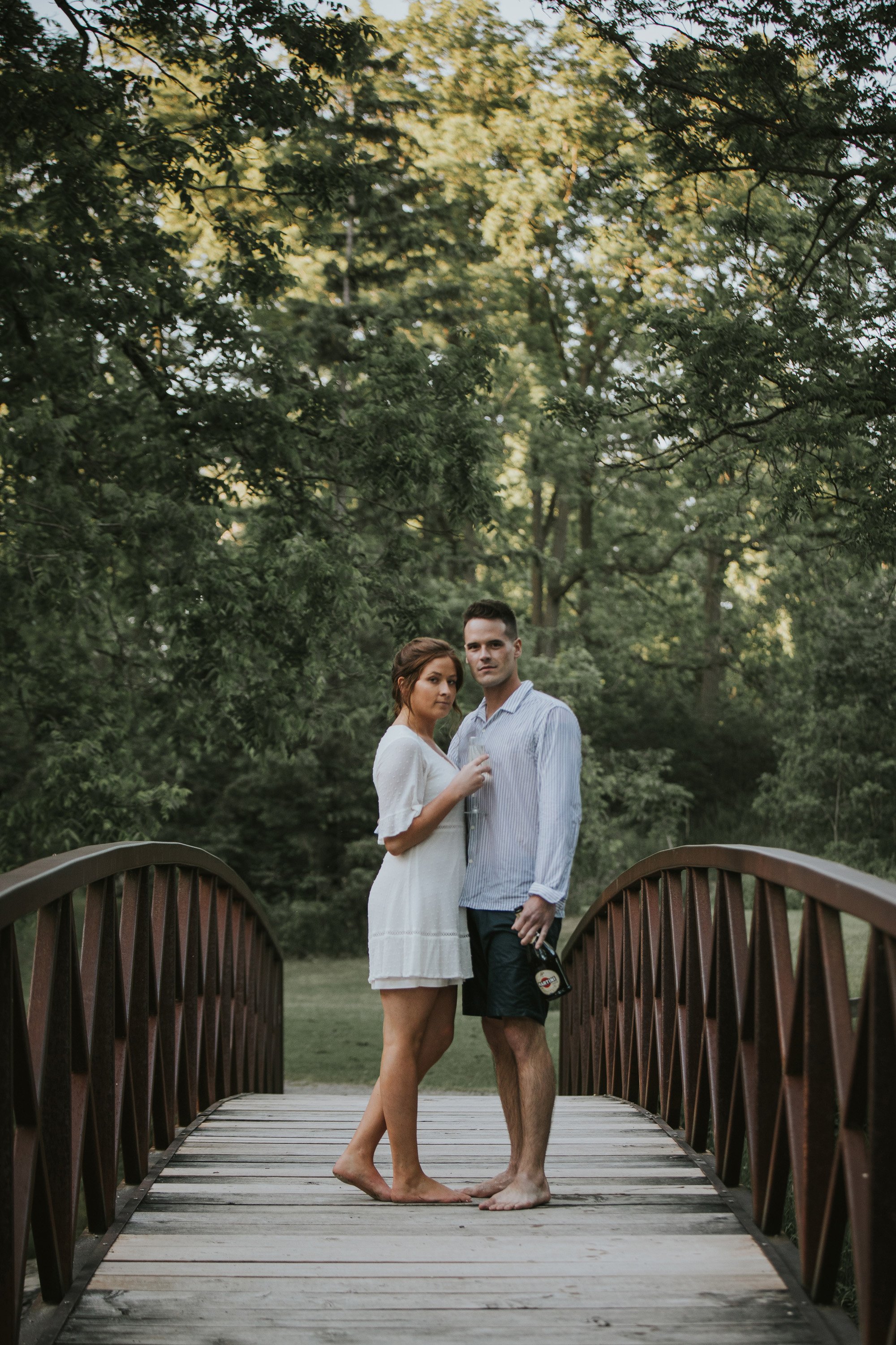 A couple stands barefoot on a wooden bridge in a forested area during their engagement session in London, Ontario. They are embracing, surrounded by lush green trees.