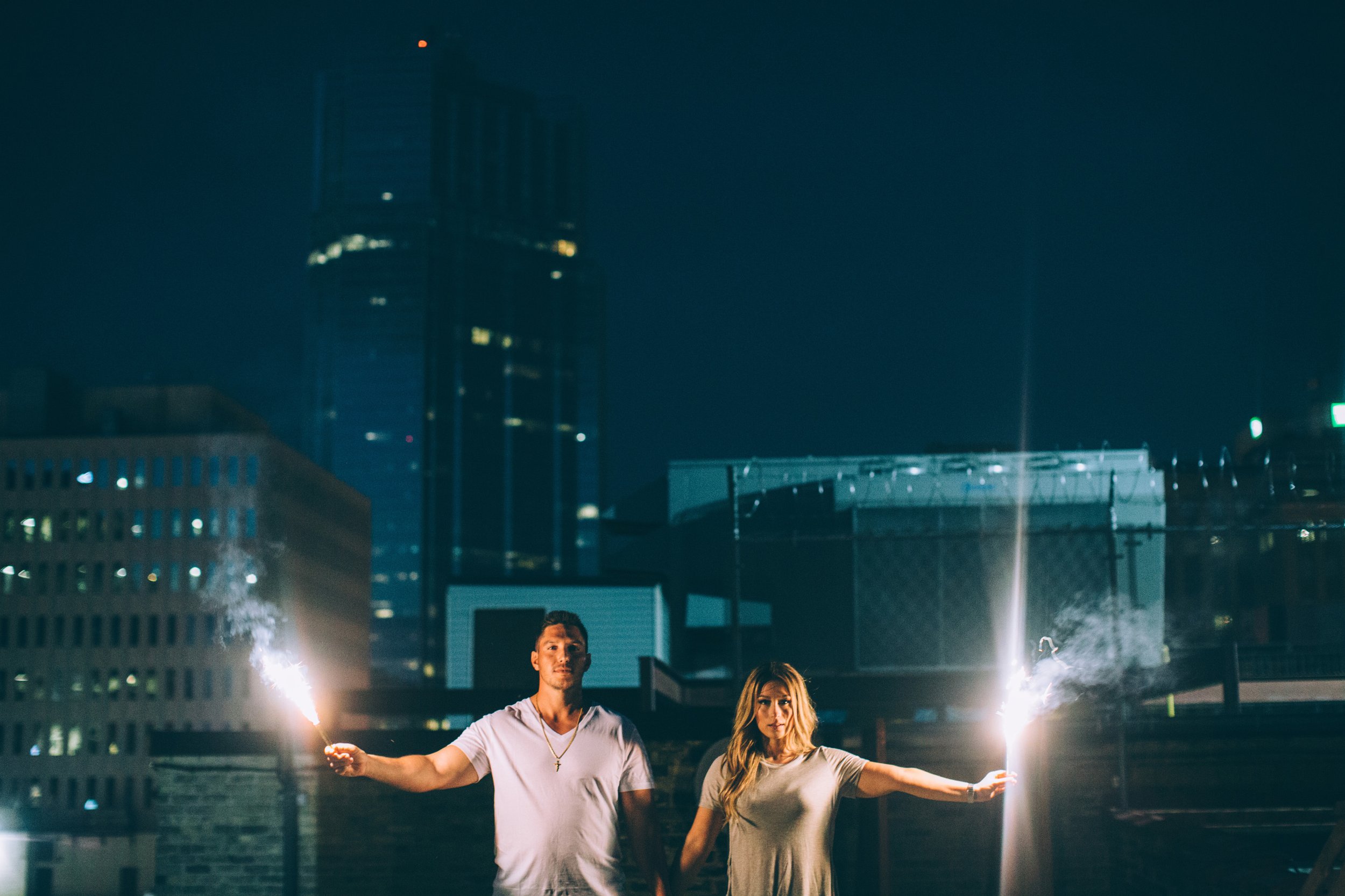 A couple holding sparklers on an urban rooftop at night, celebrating their engagement with the city skyline in the background.