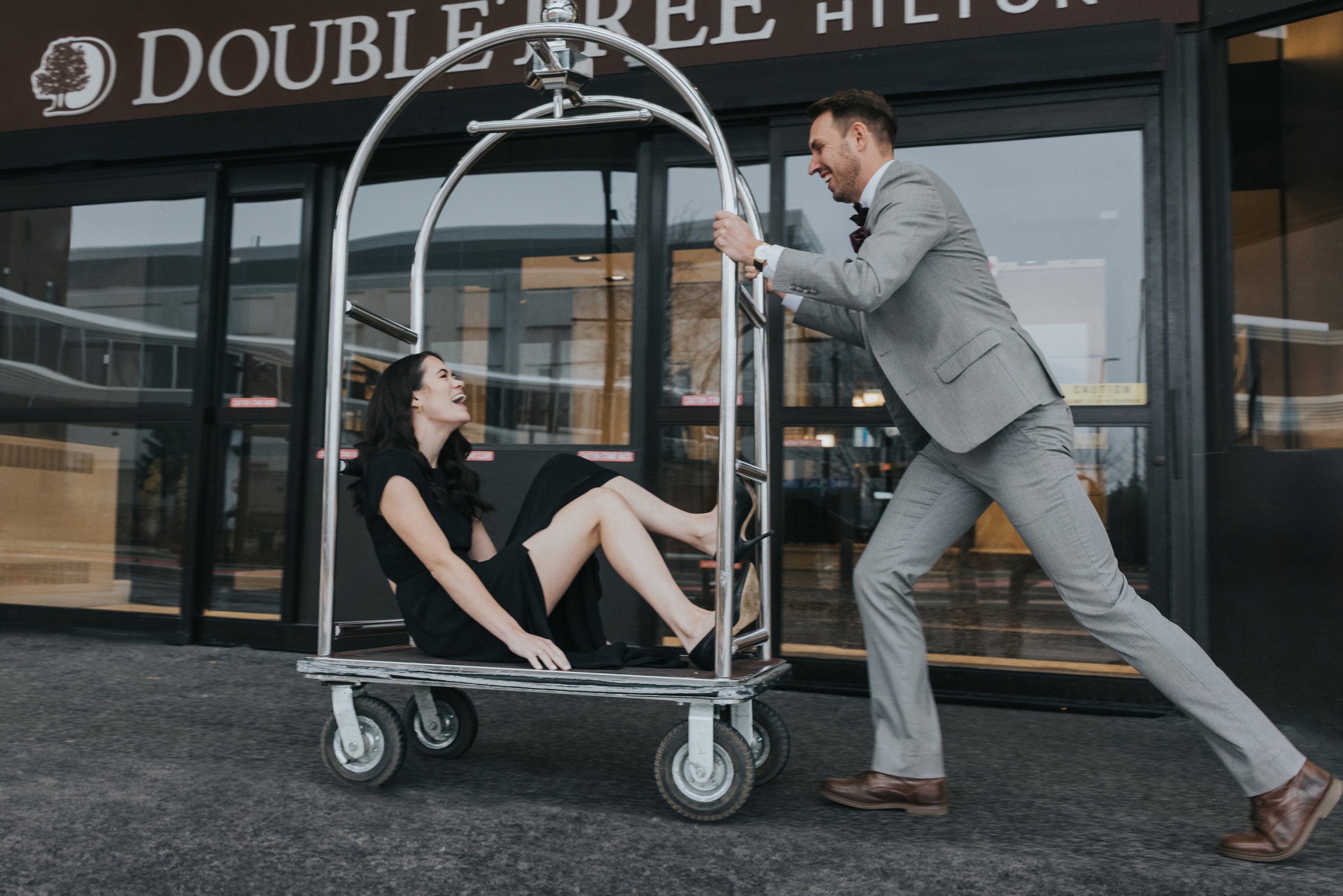 A couple enjoys a playful moment as the man pushes the woman on a hotel cart outside a DoubleTree Hilton hotel.