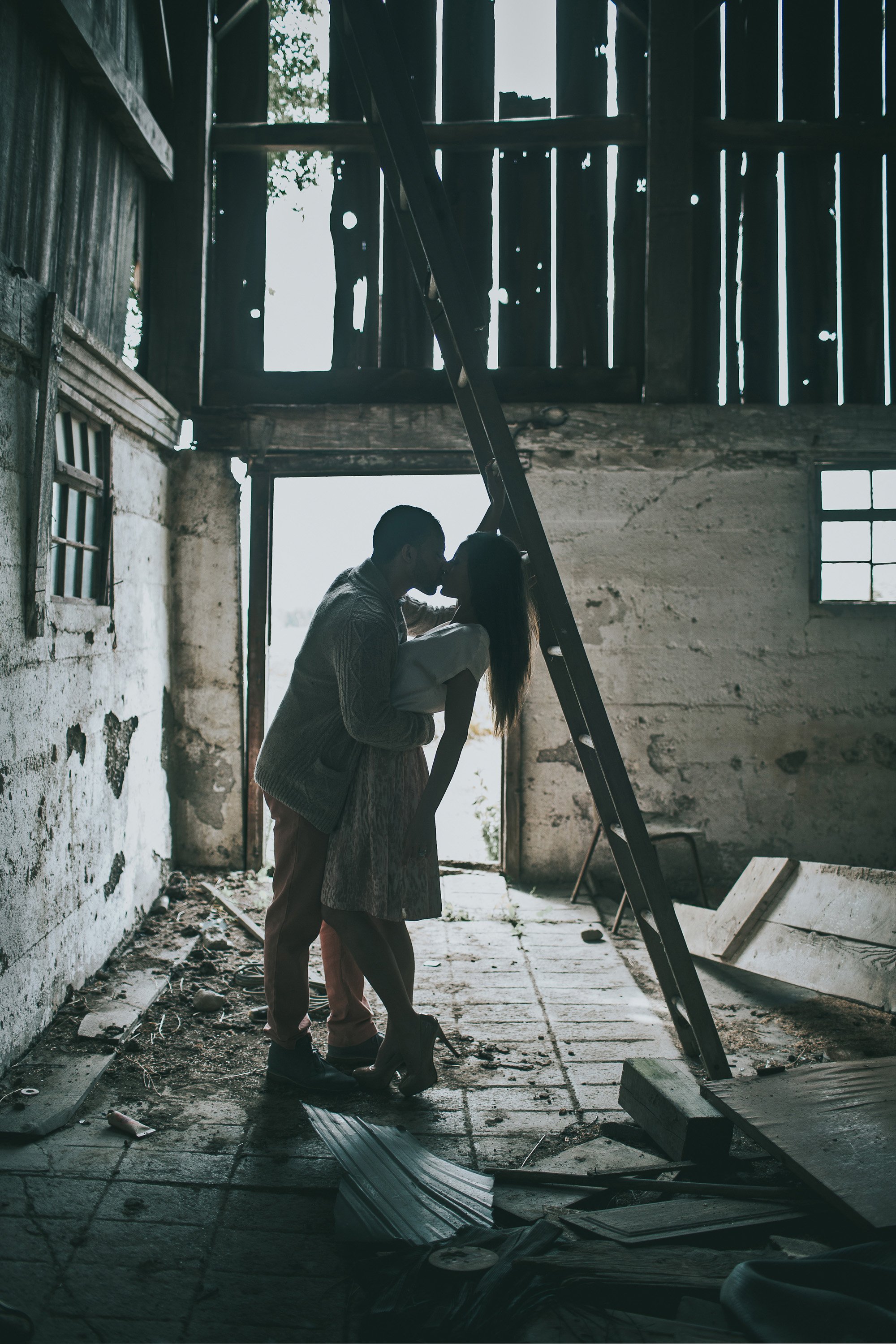 A couple shares a romantic kiss in the silhouette of a rustic barn, surrounded by weathered walls and soft, natural light.