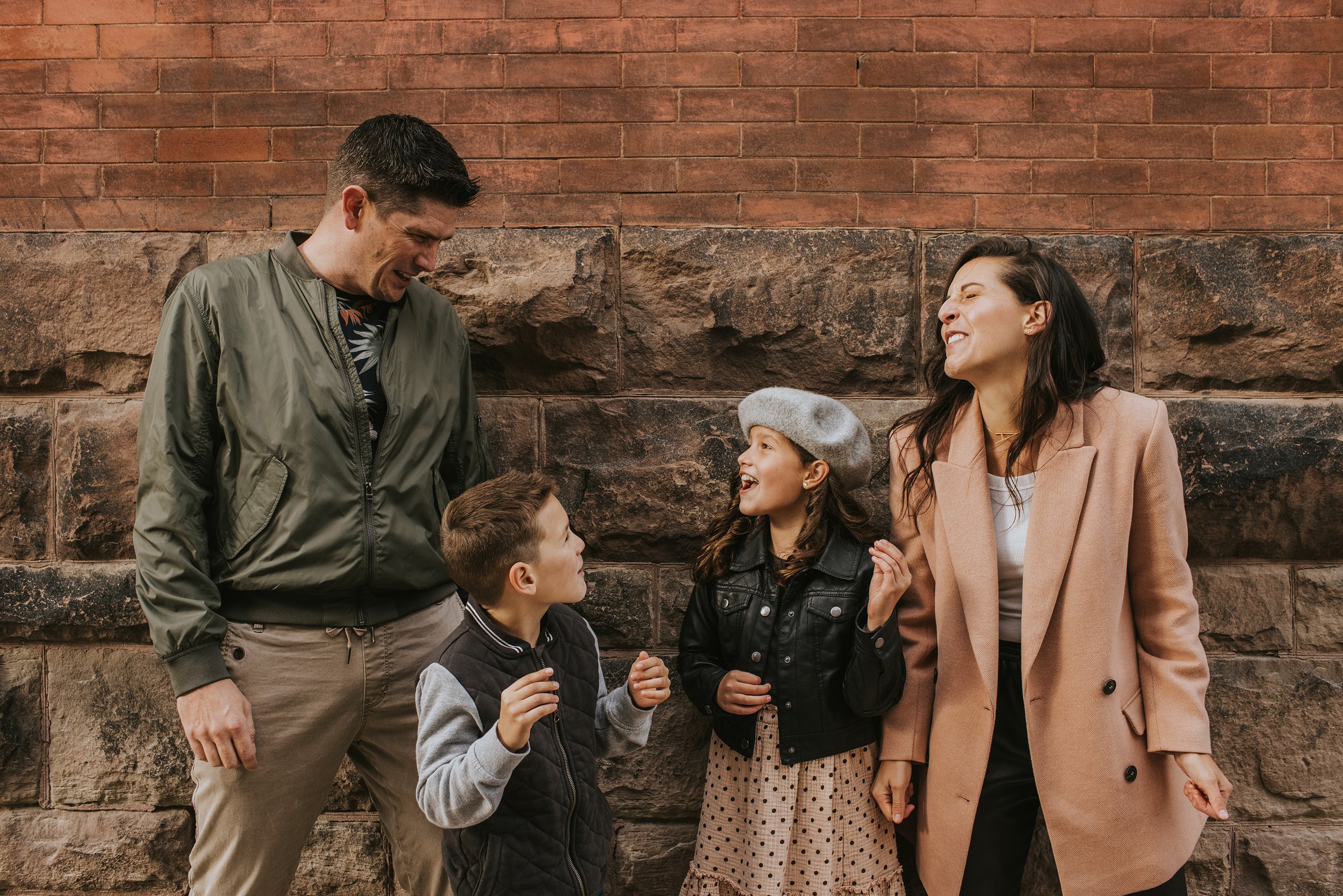 A lively urban family portrait captured against a brick wall, featuring smiles and playful interaction between parents and children.