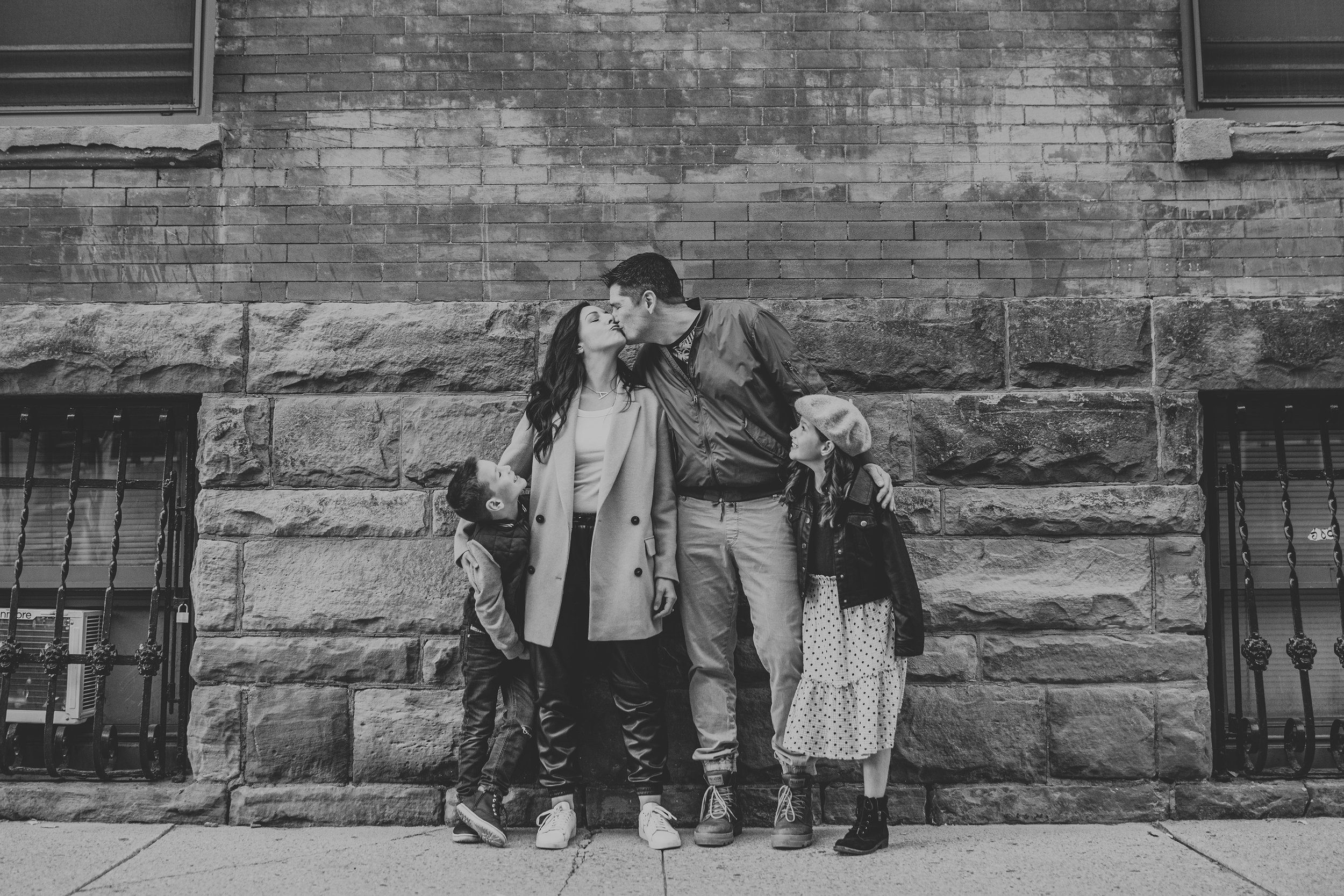 Black and white photo of a family of four sharing a kiss in front of a brick wall, with the children looking up at their parents in an urban setting.