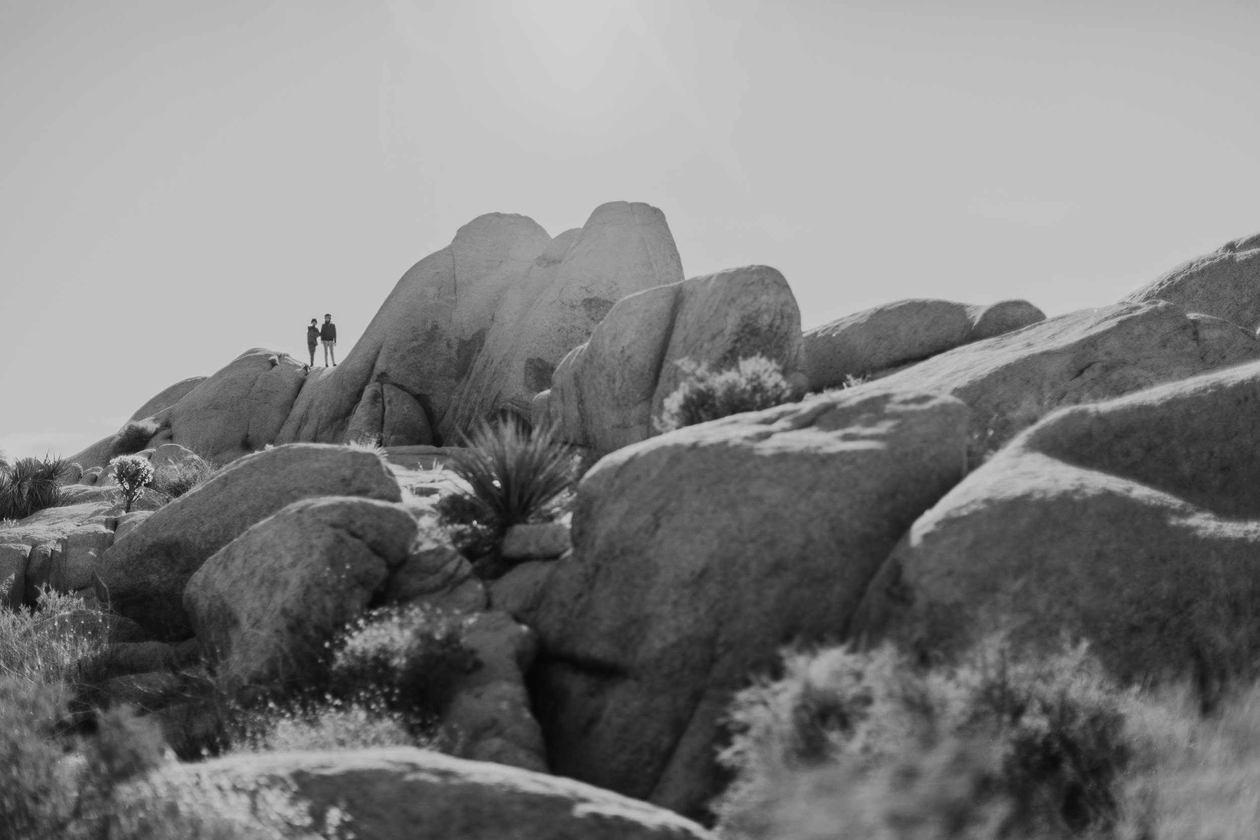 A couple standing as silhouettes on top of large rocks, set against a bright sky in a natural, adventurous landscape.