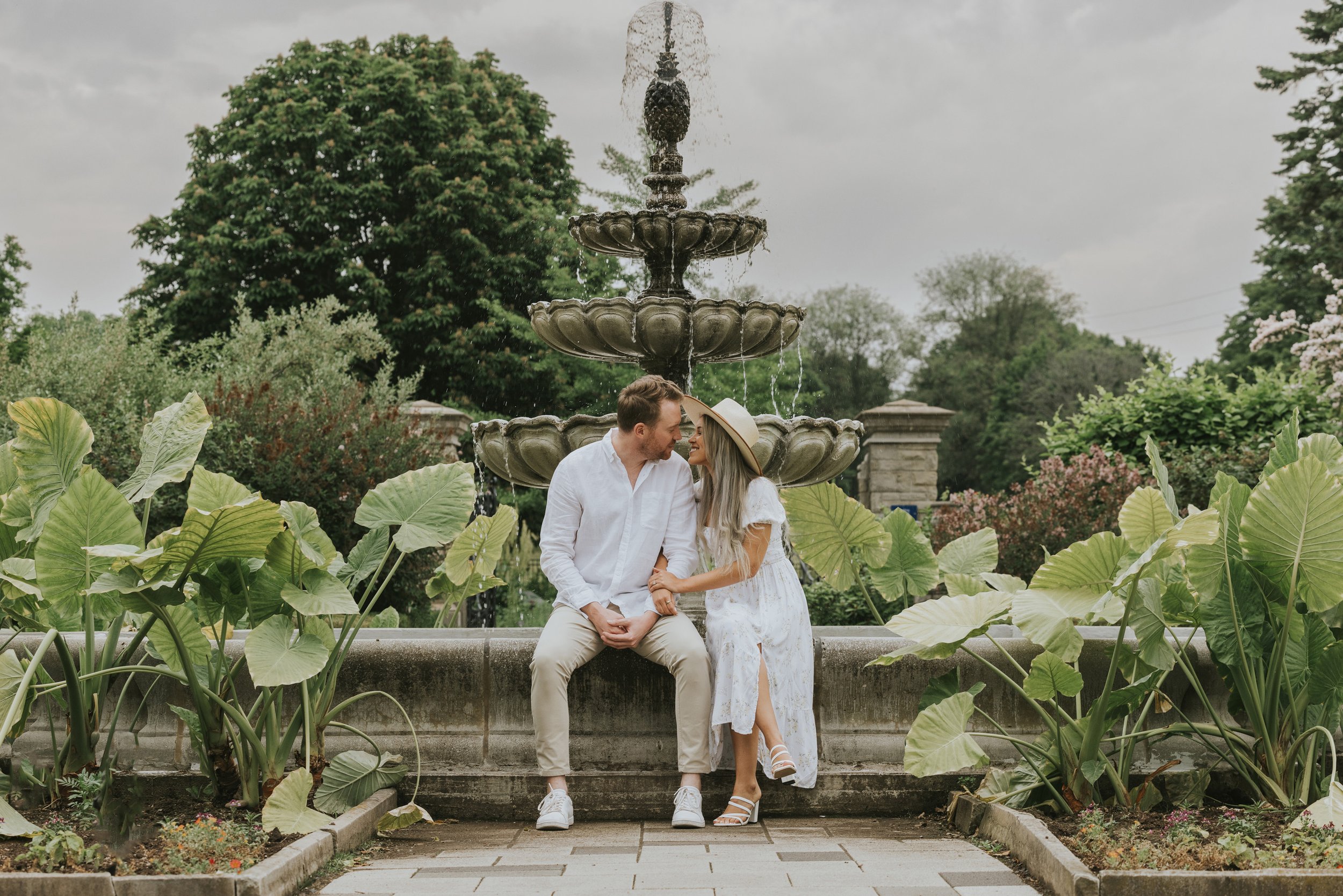 A couple sits on the edge of a beautiful garden fountain, smiling lovingly at each other, surrounded by lush greenery and large tropical leaves.