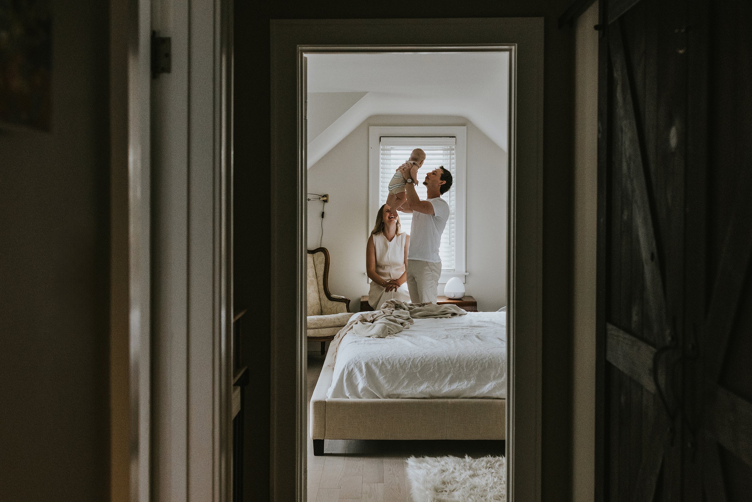 A heartwarming candid moment of a family in a cozy bedroom, with the father playfully lifting their baby into the air while the mother looks on lovingly.