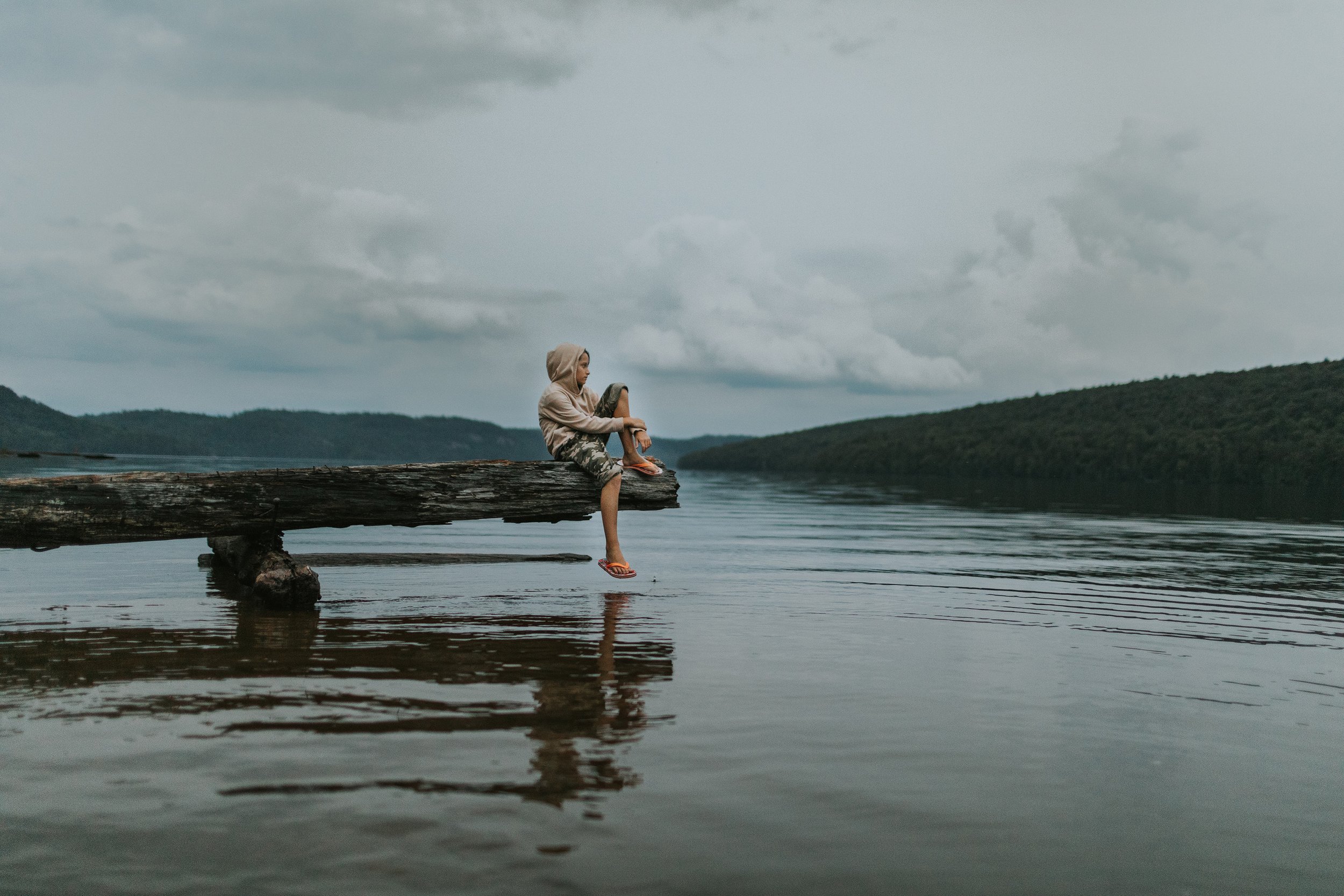 A child sitting quietly on a log by a serene lake, surrounded by nature.
