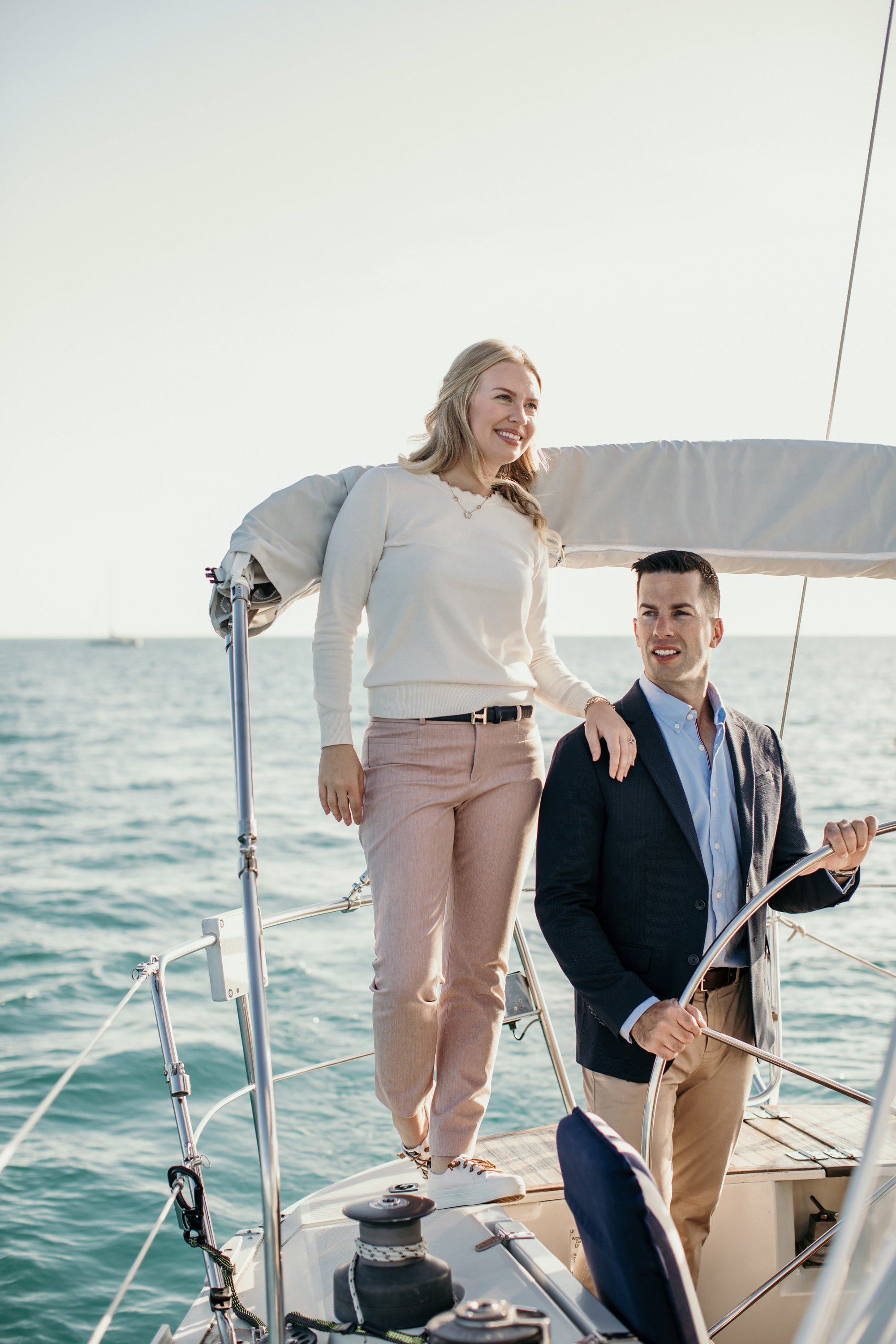 A couple enjoys a luxurious engagement photo session aboard a yacht, with the man steering the boat and the woman standing beside him, both looking out over the serene waters of Ontario.