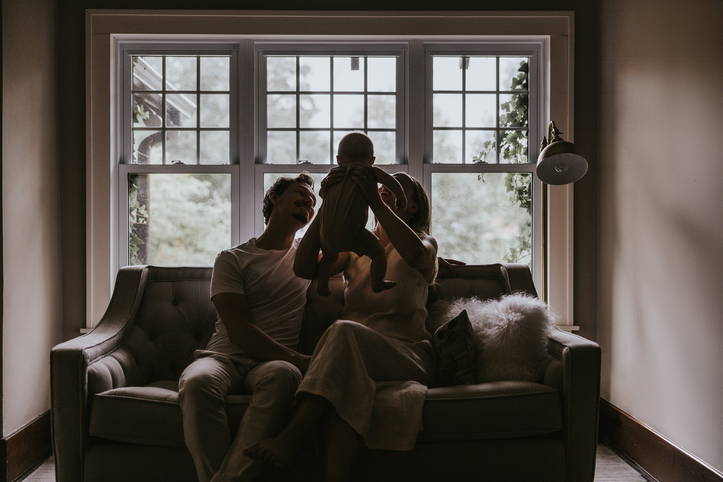 A touching silhouette of a family sitting on a couch, holding their baby up to the light of a large window, capturing an intimate and serene moment.