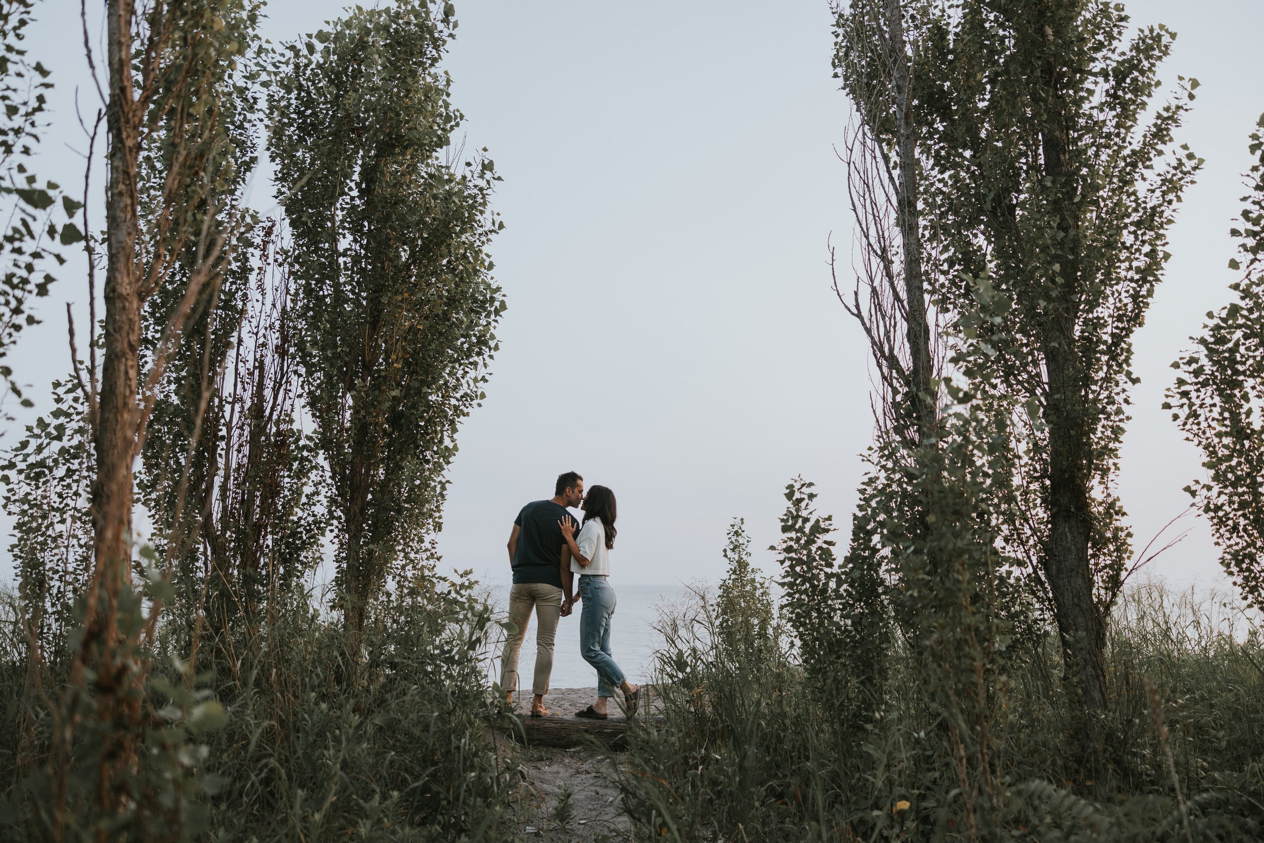 A couple shares a kiss during a nature walk, framed by tall trees and lush greenery, creating a romantic and serene atmosphere.