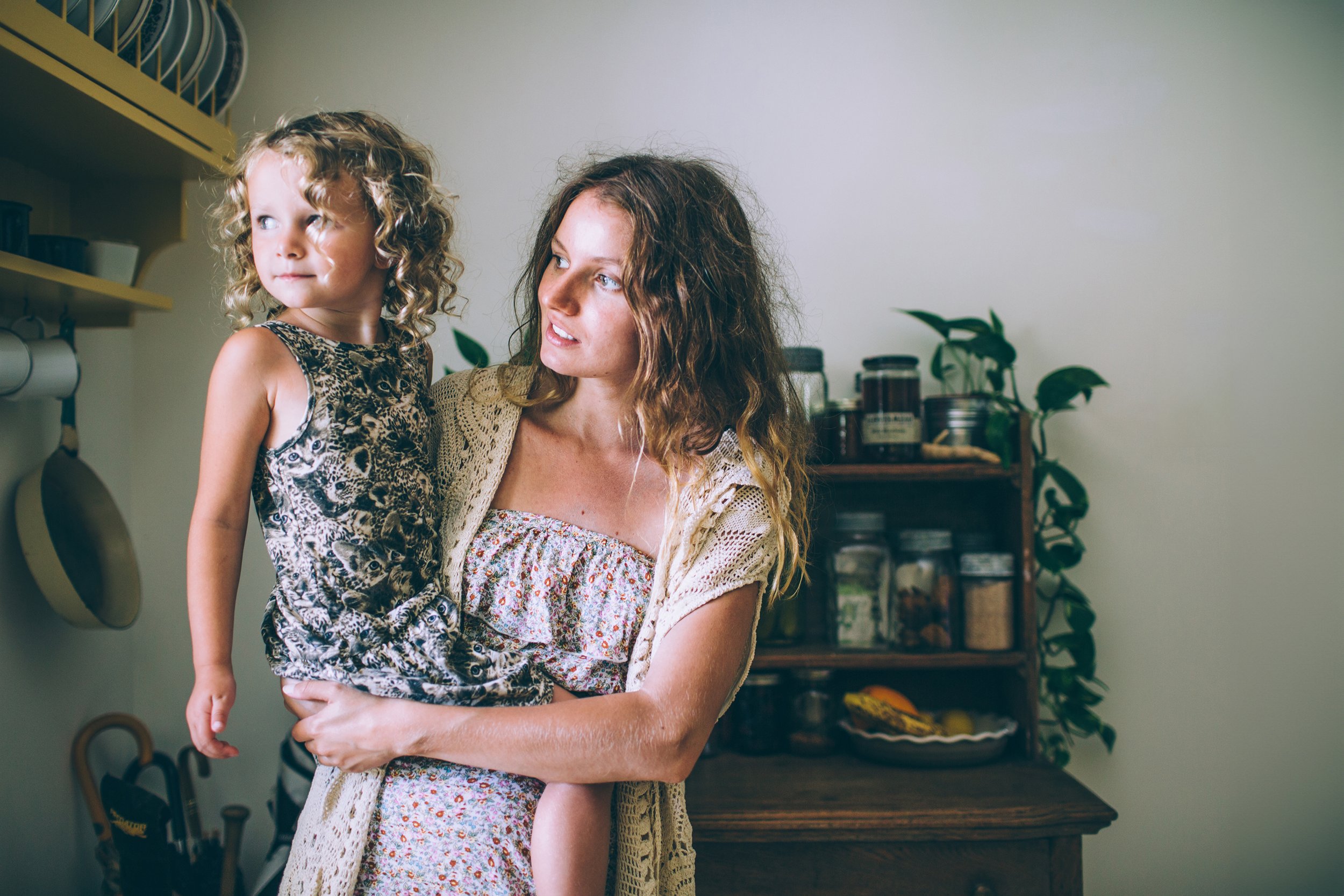 A candid portrait of a mother holding her child in a vintage-style kitchen, both gazing thoughtfully out the window.