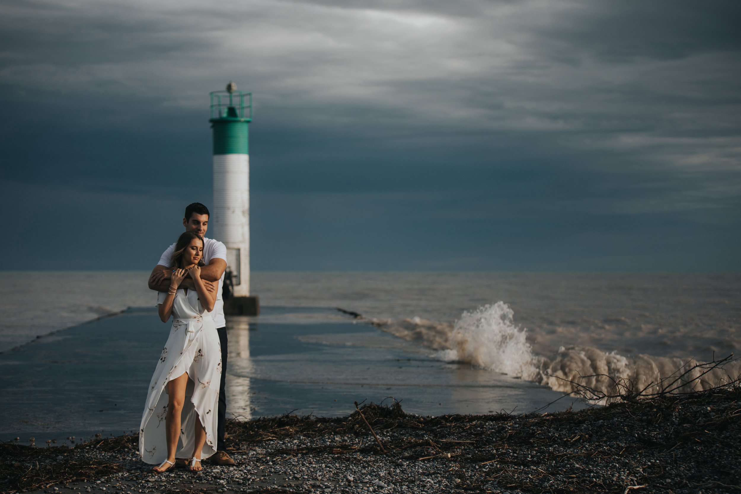 A couple embraces on a pier with a lighthouse in the background, under a dramatic sky with waves crashing nearby.