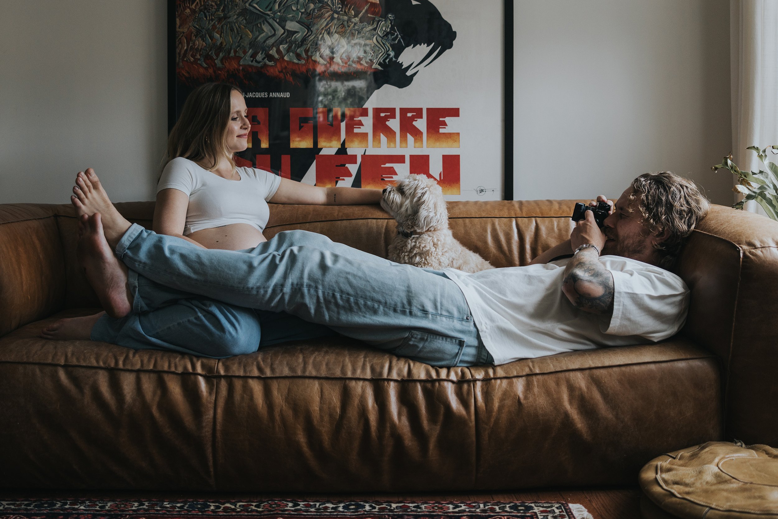 A relaxed couple lounging on a couch with their dog during a casual home pregnancy session, capturing a cozy and intimate atmosphere.
