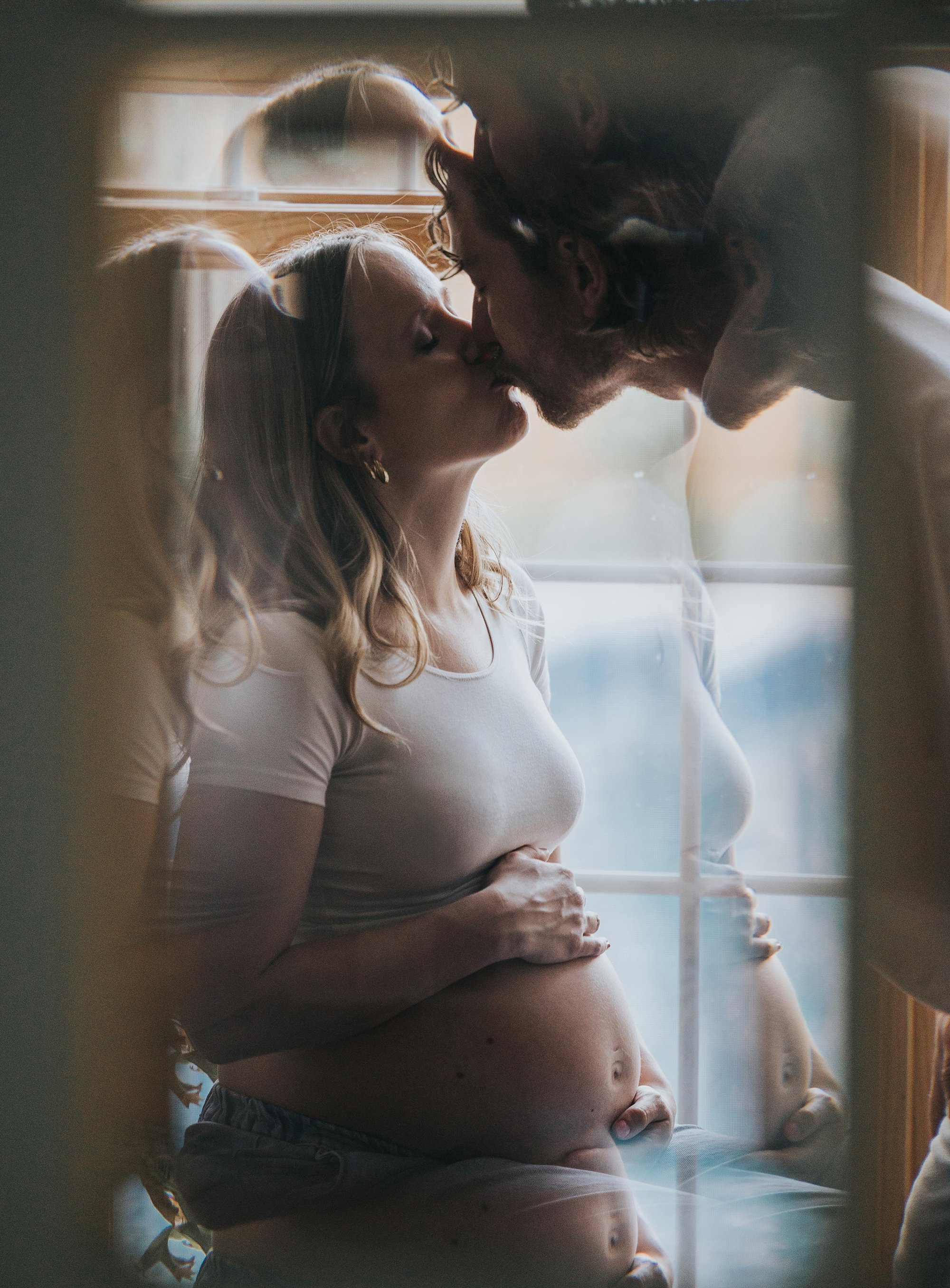 A creative portrait capturing a pregnant couple kissing with their reflection mirrored in a window.