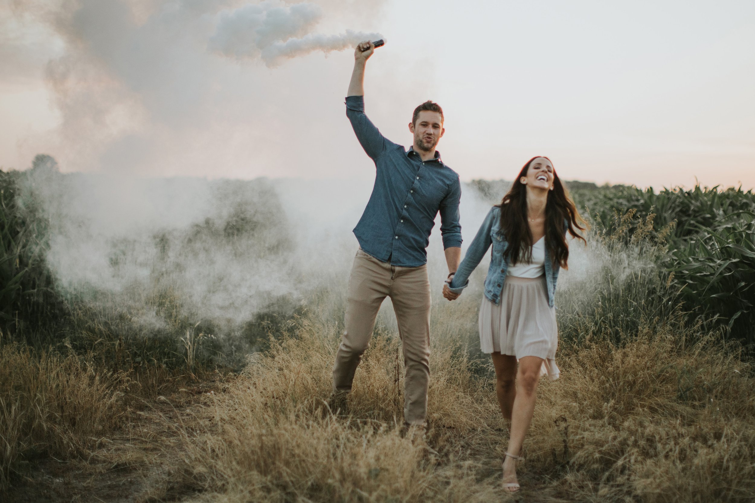 A couple walks through a field with a smoke bomb, their adventurous spirit and playful energy captured in this creative engagement photograph.