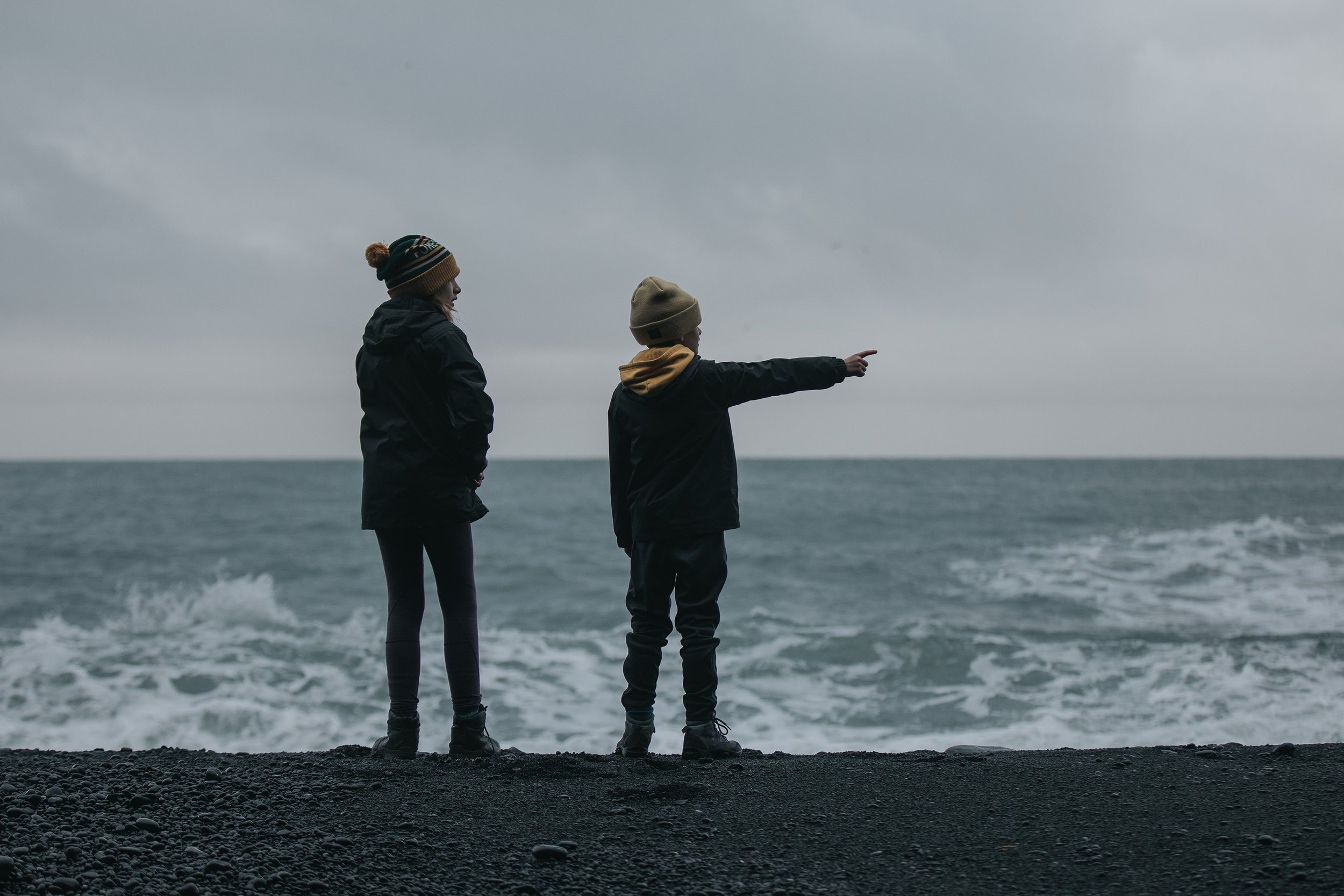 Two children, dressed warmly, stand on a rocky beach, with one pointing towards the ocean under a cloudy sky.