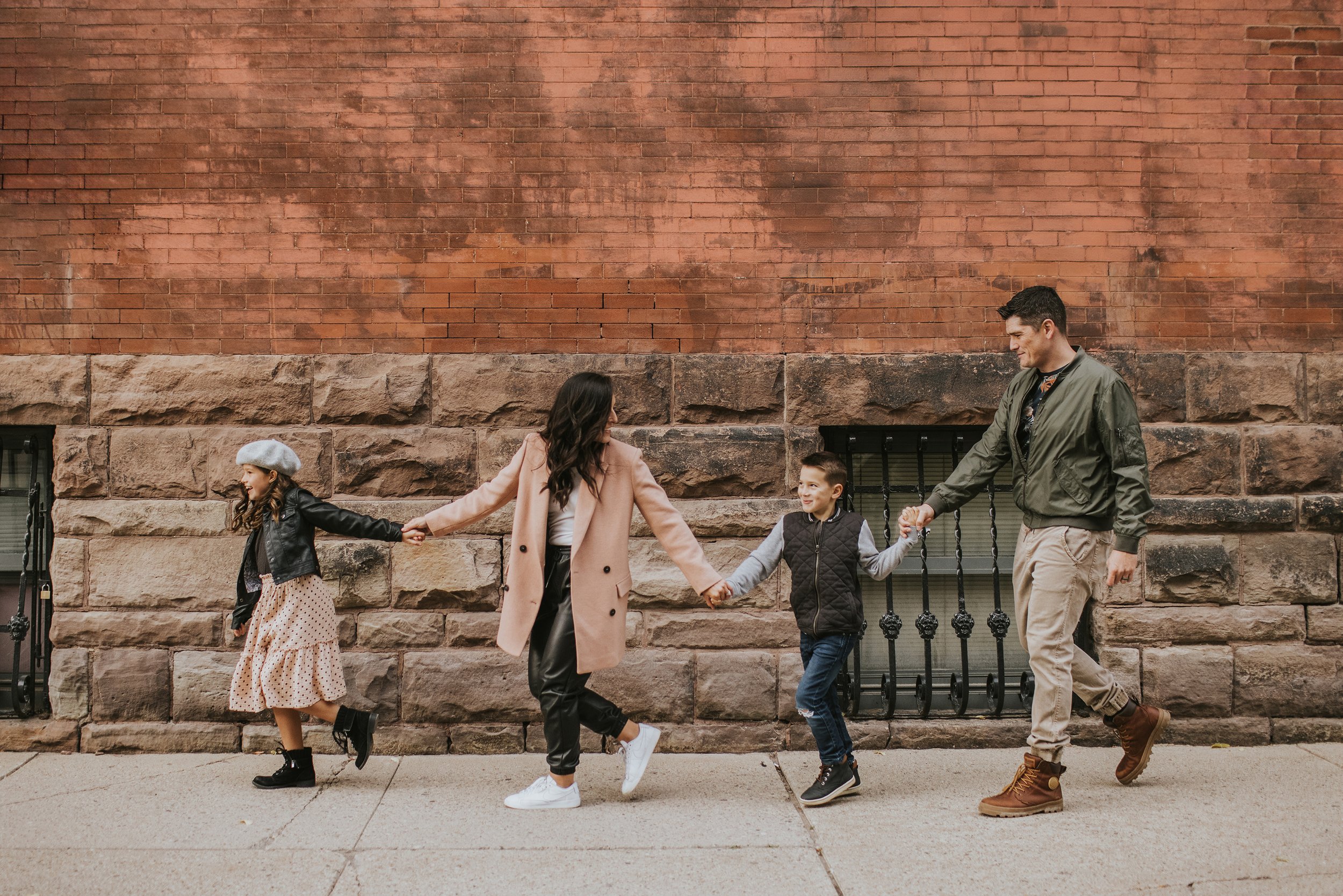 A family of four walking hand-in-hand along a brick wall, smiling and enjoying a casual urban stroll together.