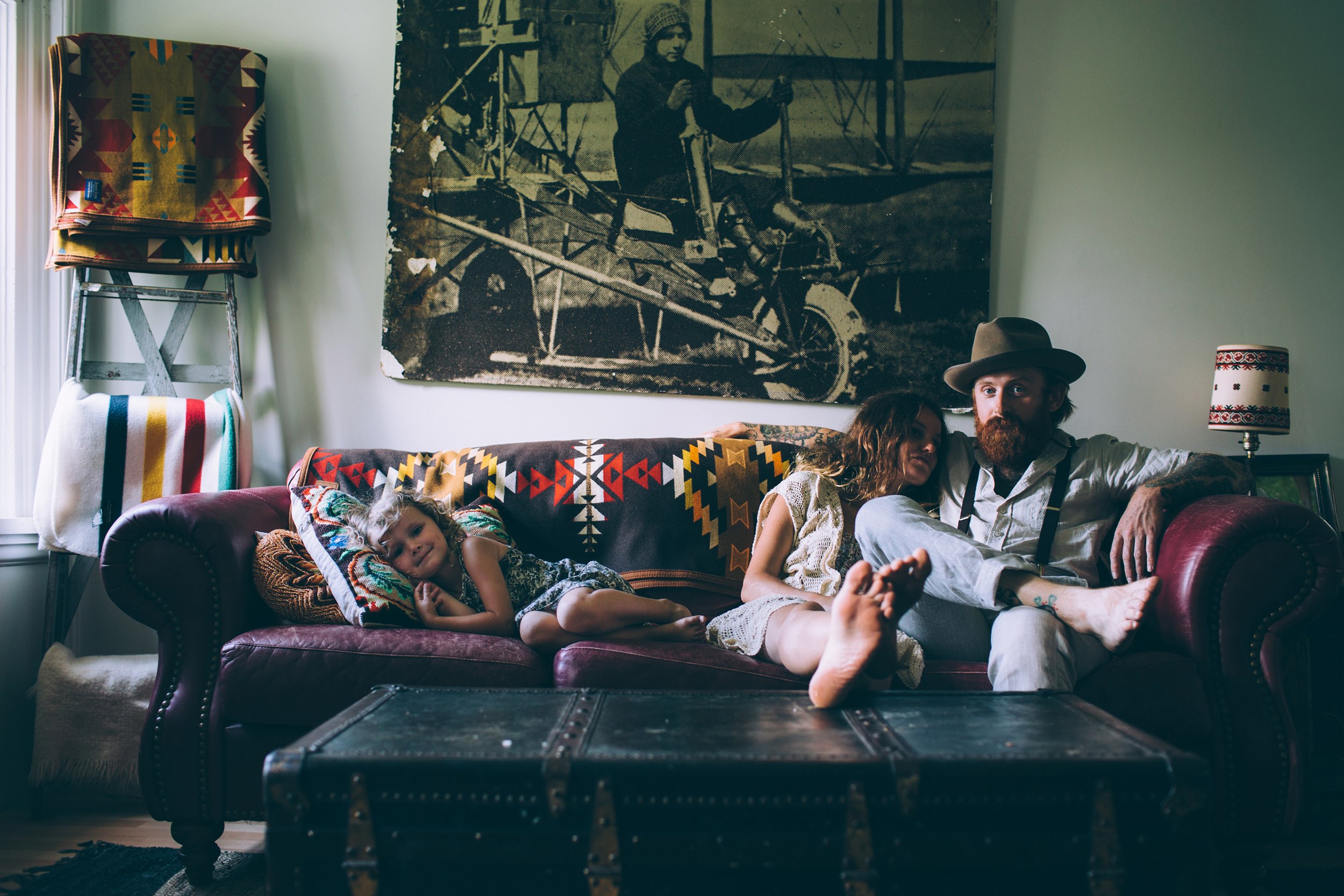 A relaxed, cozy family scene inside a vintage home. The family sits together on a couch, surrounded by rustic decor, including a vintage poster on the wall. The atmosphere is laid-back, with a sense of warmth and togetherness.