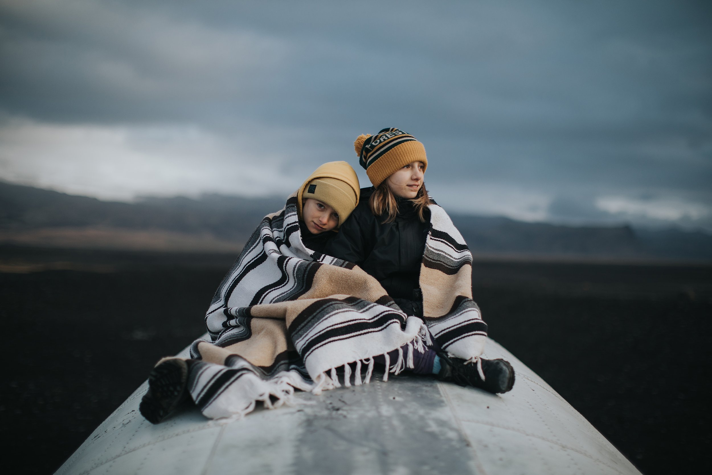 Two children cuddled together under a blanket, sitting outdoors in a wide-open landscape, wearing warm hats as they enjoy the tranquil, chilly environment.
