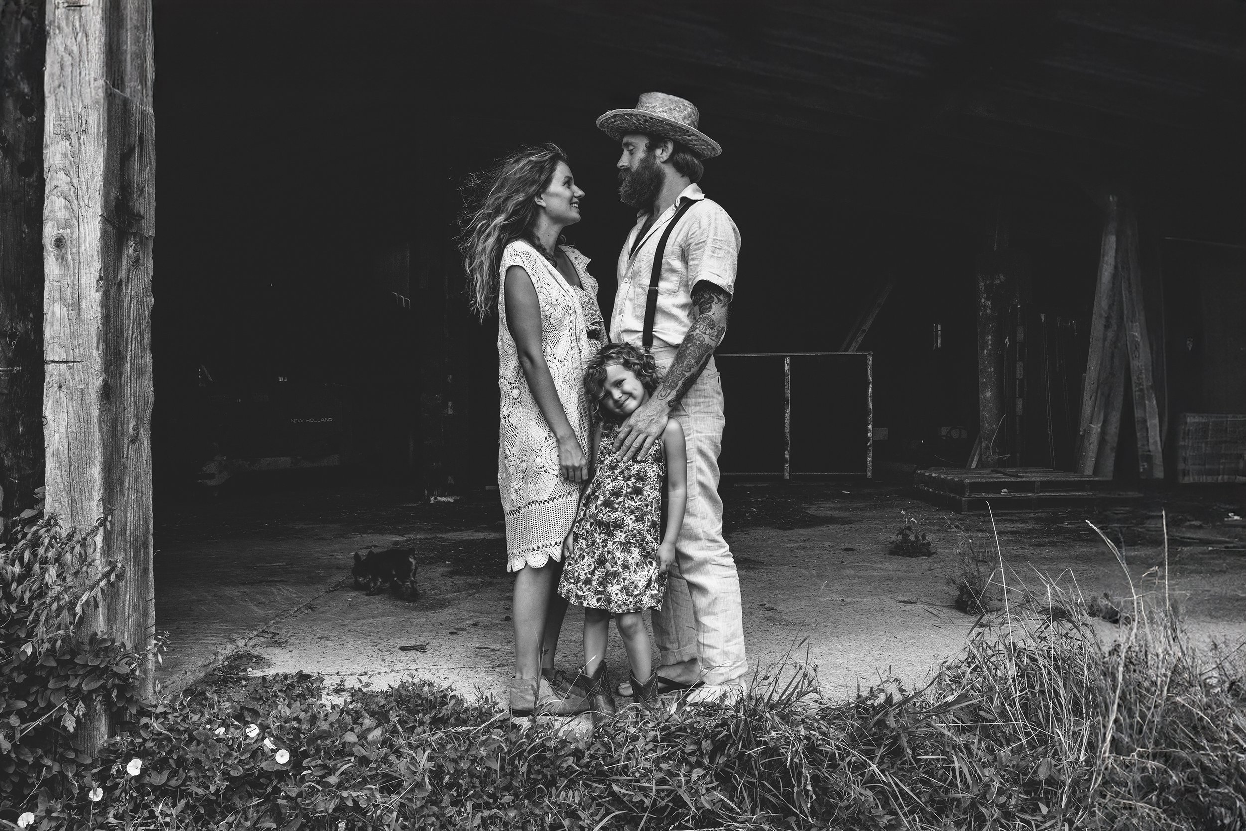 A black-and-white rustic family portrait featuring a couple and their young daughter standing together in front of a barn.