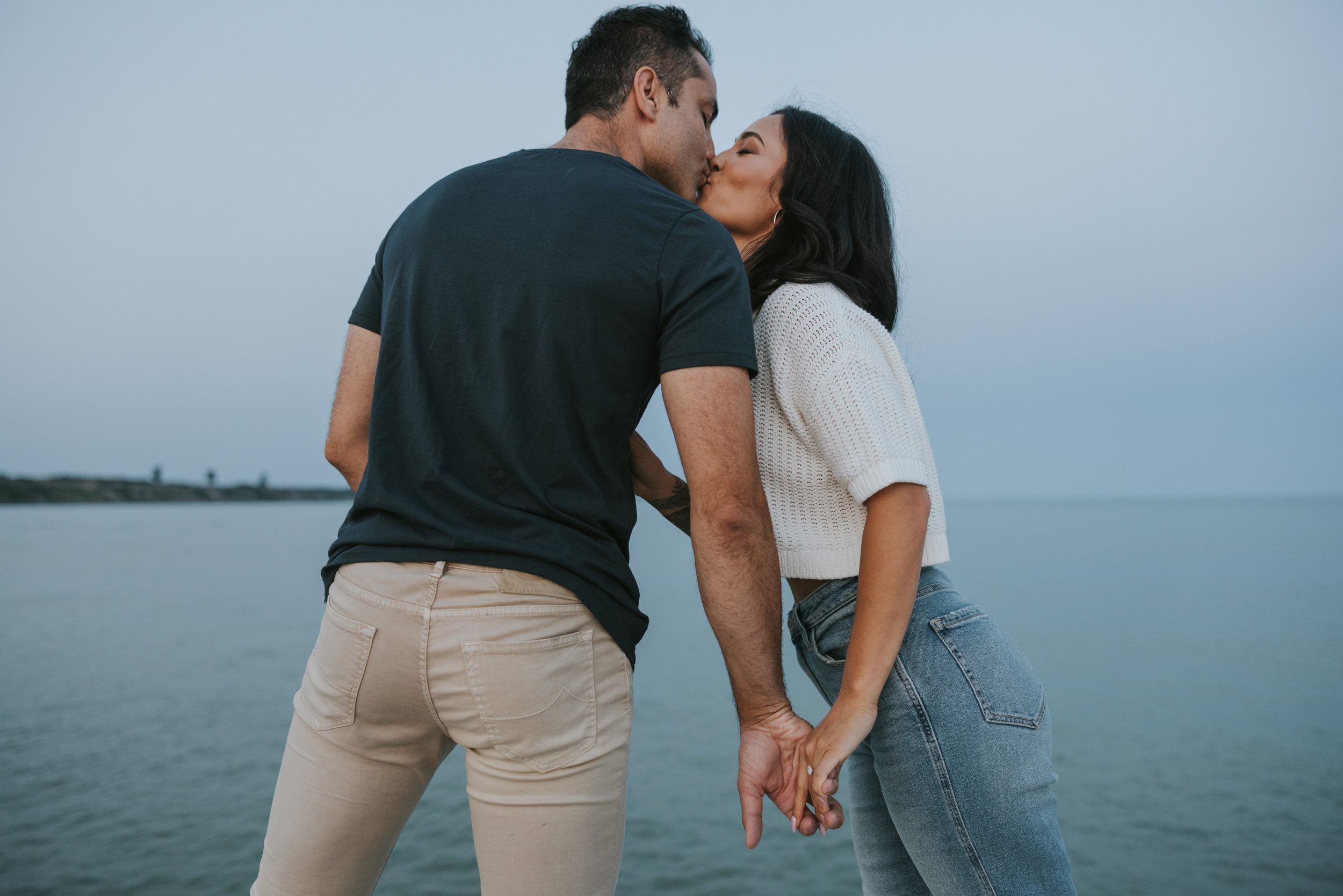 A couple shares a tender kiss by the beach, their hands intertwined. The intimate moment captures the serene and romantic connection they share with the natural surroundings of the ocean.