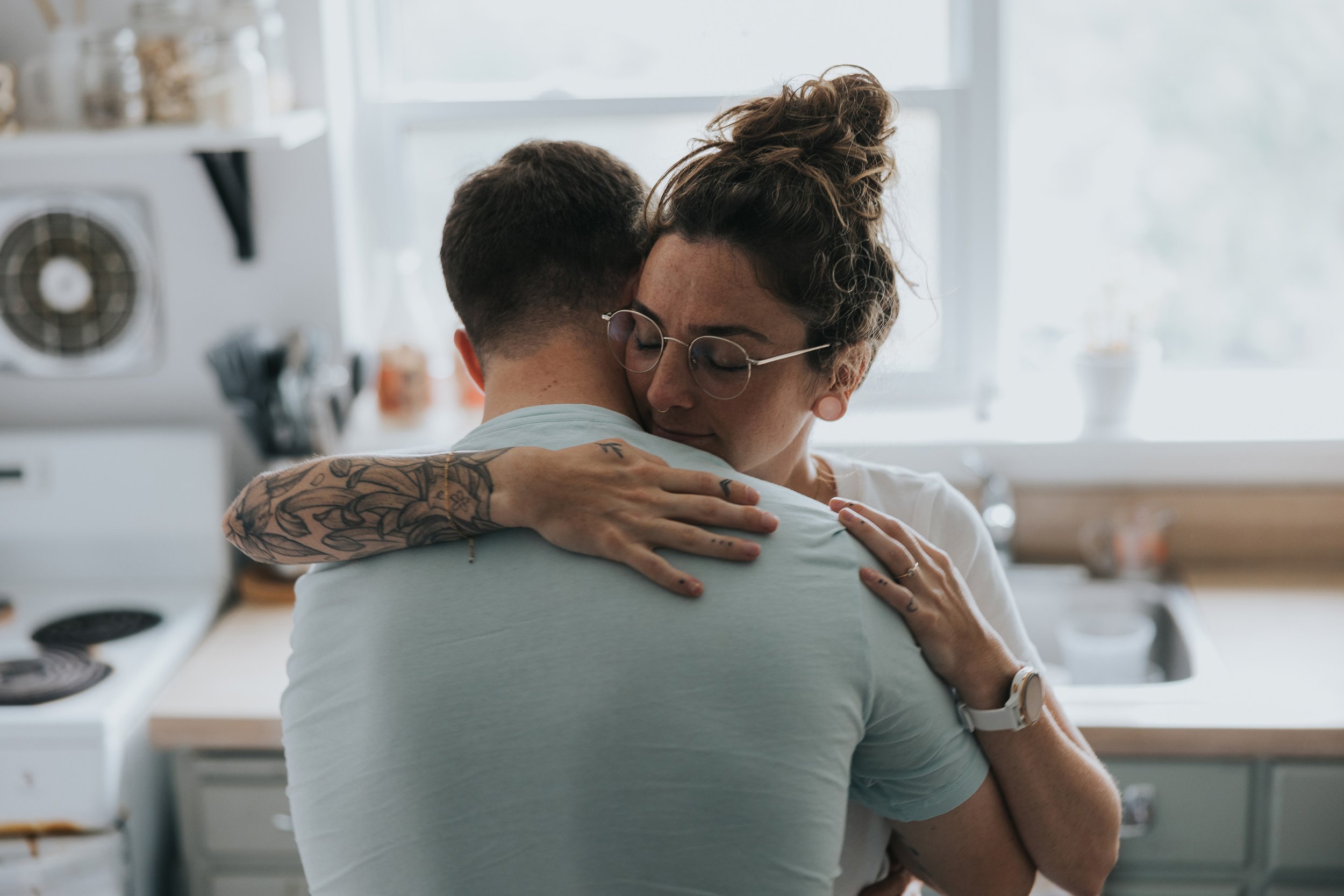 A couple shares a warm embrace in their kitchen during the morning, with sunlight filtering through the window, creating a cozy atmosphere.