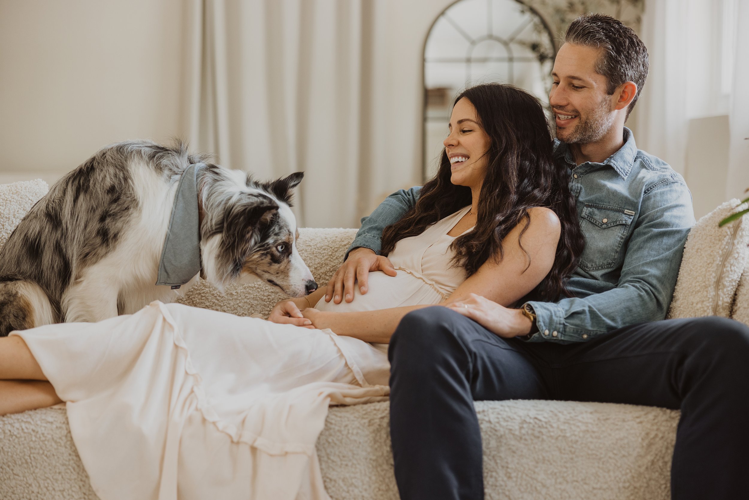 A happy couple sitting on a couch with their dog, smiling and enjoying a tender moment during a maternity photo session.