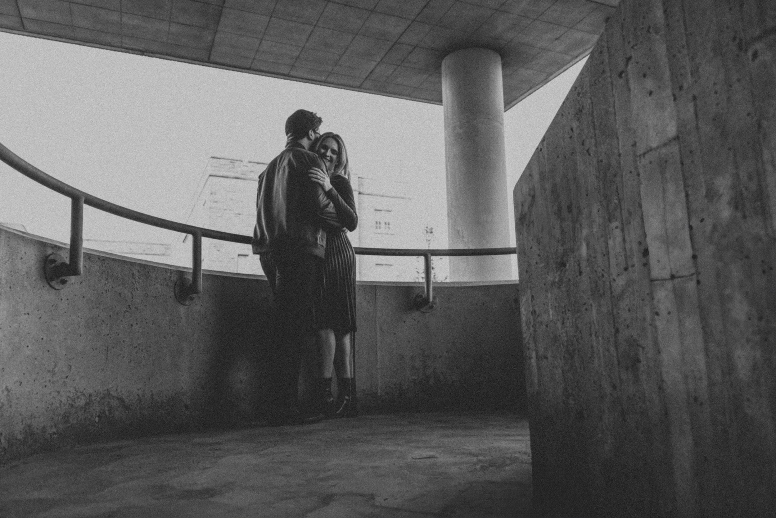 A cinematic black-and-white photograph of a couple standing in an urban architectural setting, emphasizing the strong contrasts and lines of the structure.