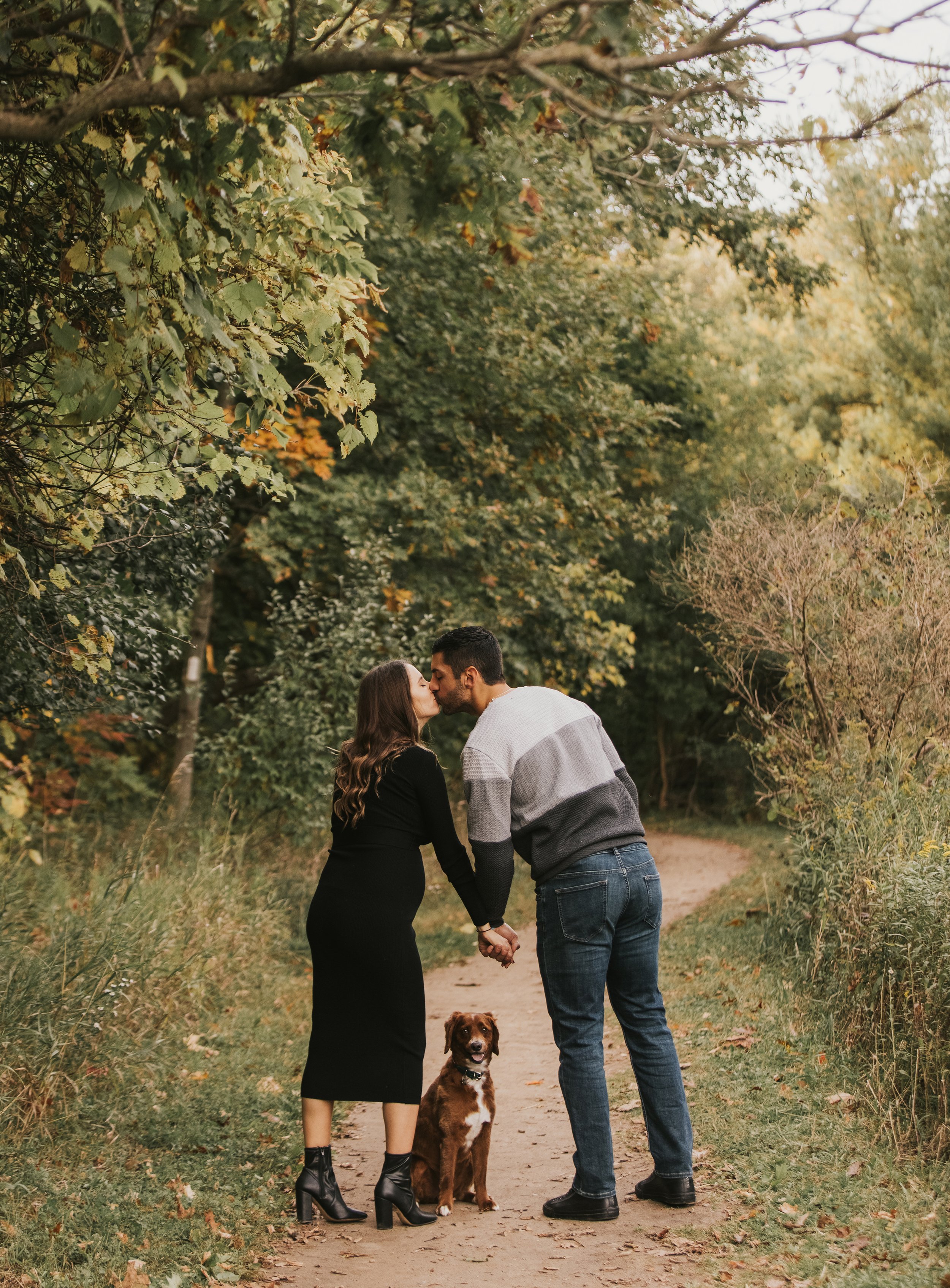 A couple walking hand-in-hand along a woodland path, sharing a kiss as their dog sits between them.