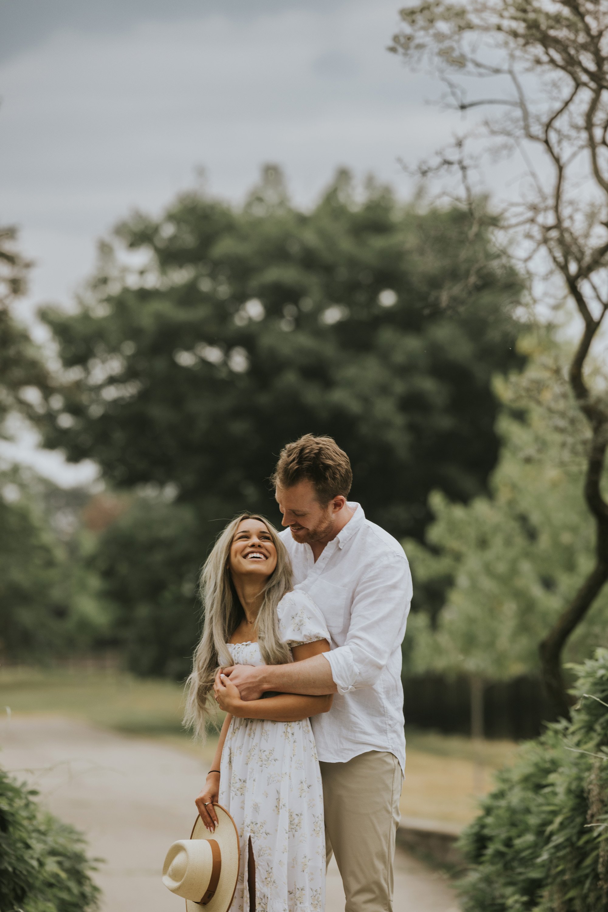 Couple embracing and smiling at each other while standing on a pathway surrounded by lush greenery on a summer day.