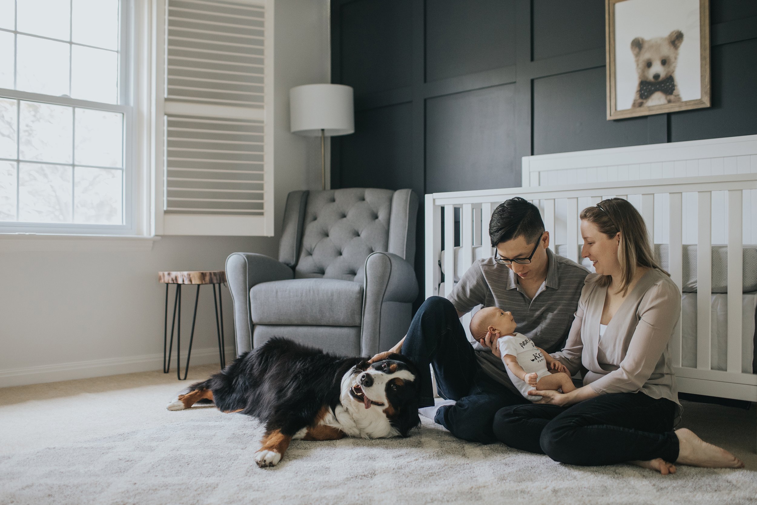A tender family moment in a nursery with parents sitting on the floor, holding their newborn baby, while a large dog lies contentedly beside them.