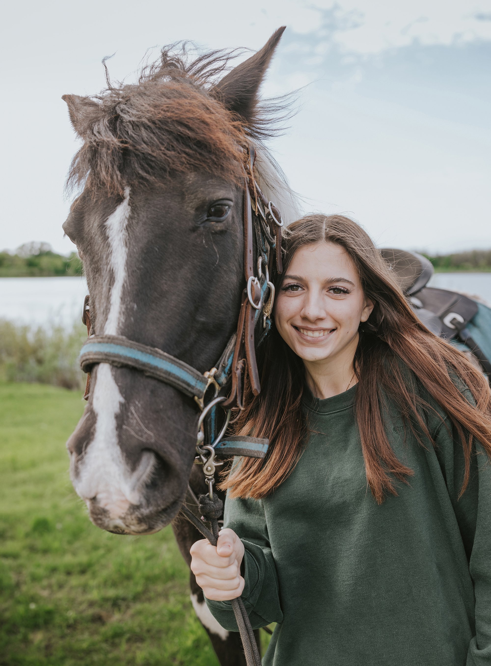 A young girl smiling brightly while standing next to a brown horse in a grassy field.
