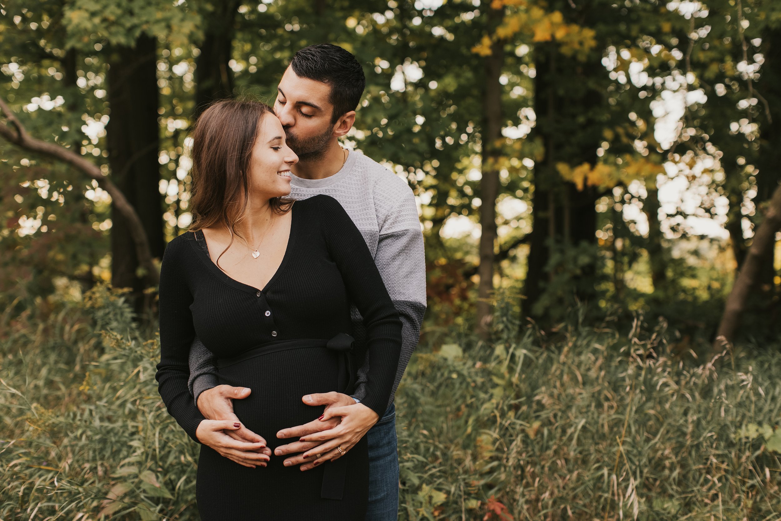 A romantic outdoor portrait of a couple sharing a kiss, with the husband embracing his pregnant wife in a lush natural setting.