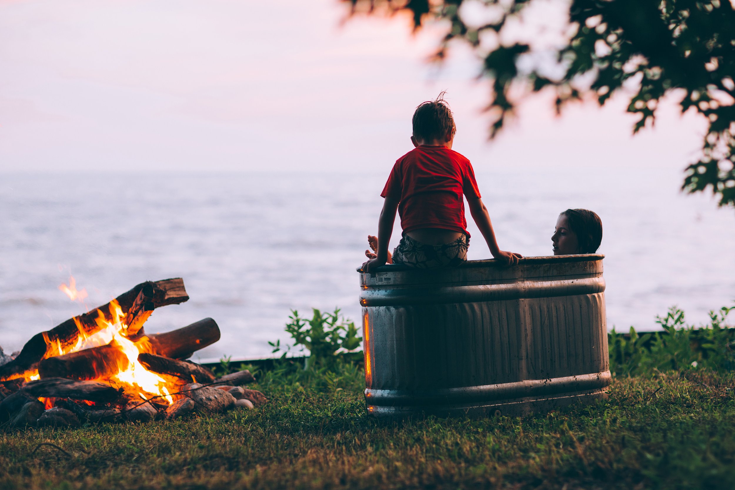 Two children enjoying a warm evening by a lakeside campfire, one sitting in a tub, capturing the essence of carefree outdoor play.