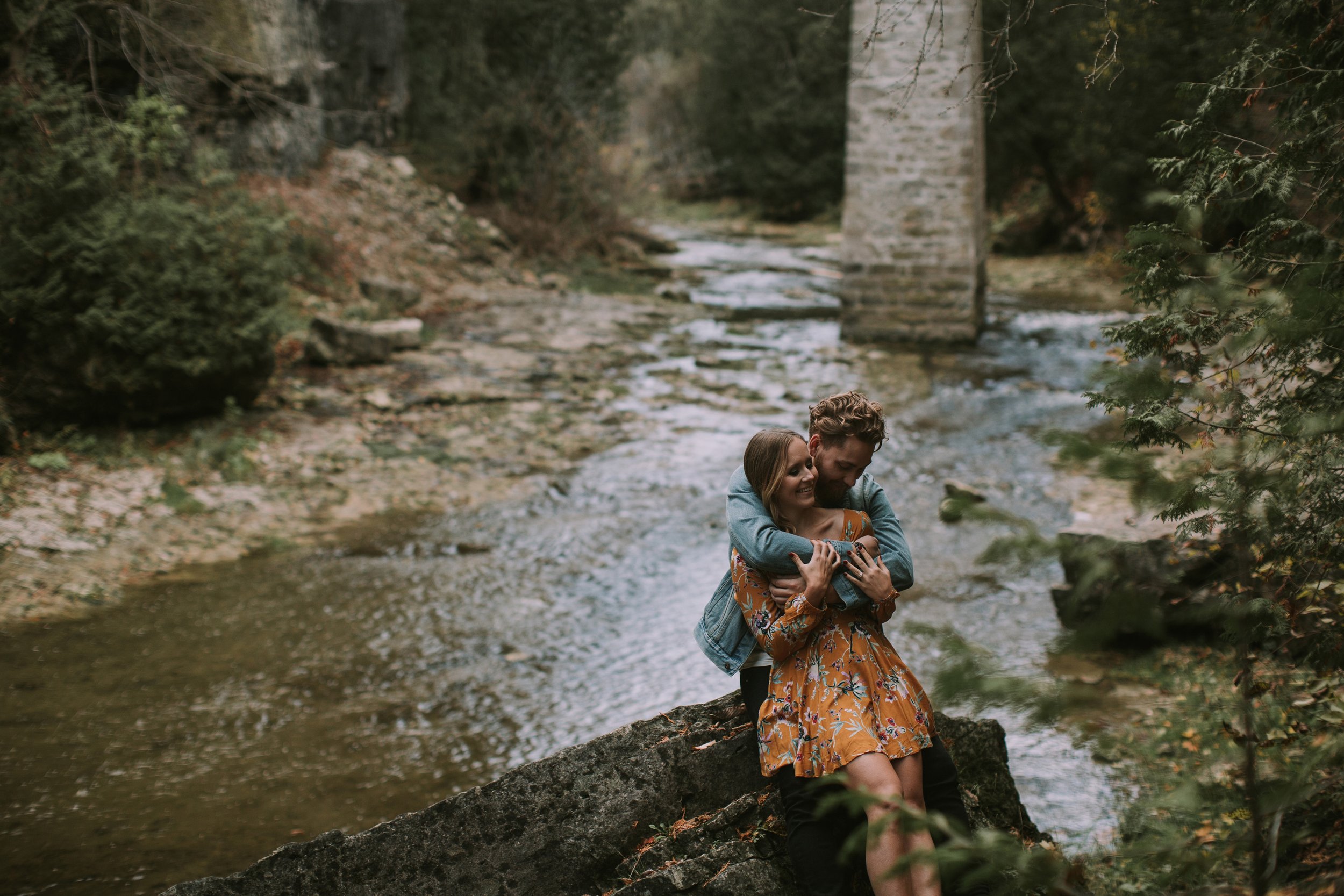 A couple hugs by a river during the fall, surrounded by colourful leaves and serene nature, capturing a moment of warmth and love.