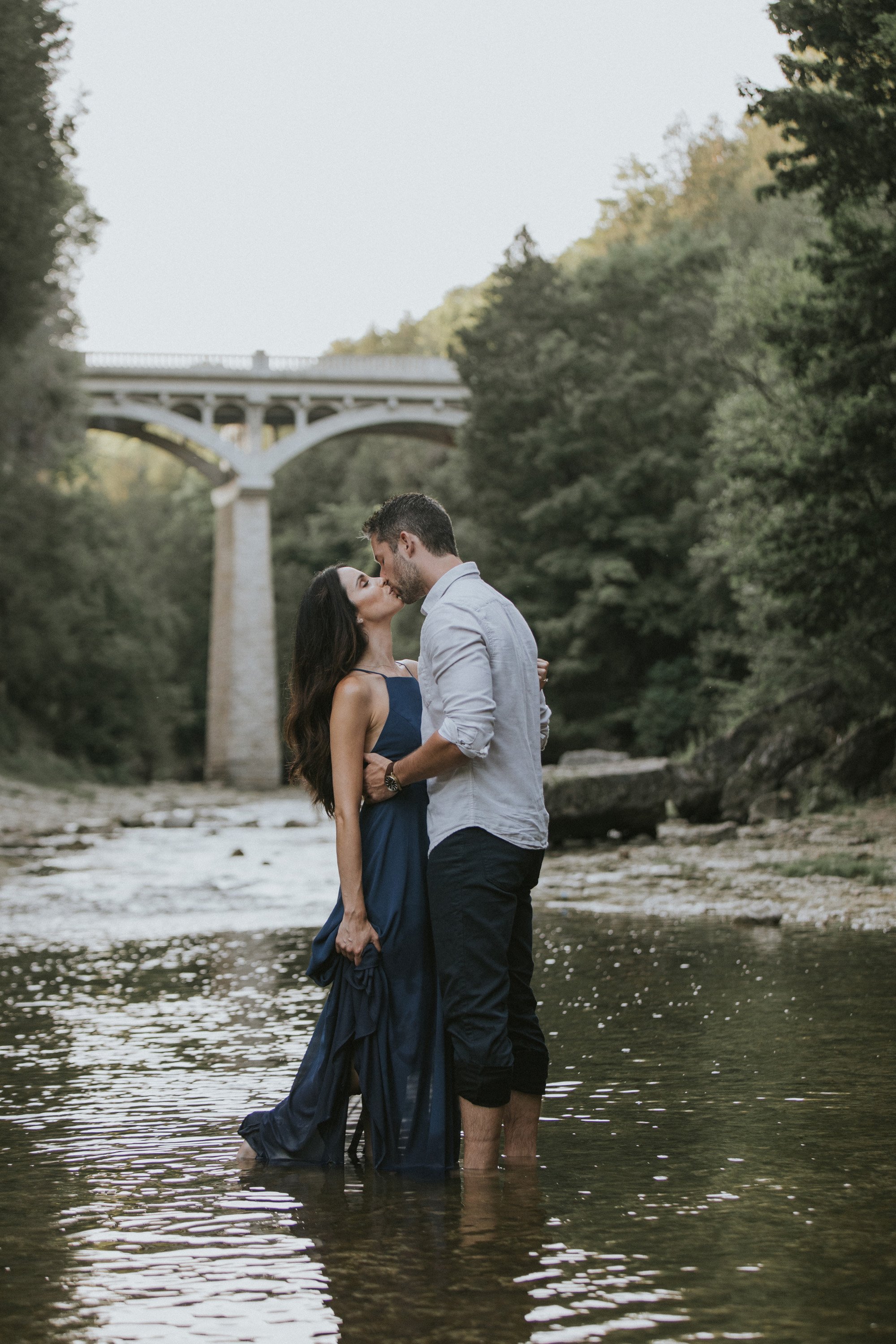 A couple sharing a romantic kiss while standing in a shallow river, with the Elora Gorge bridge in the background.
