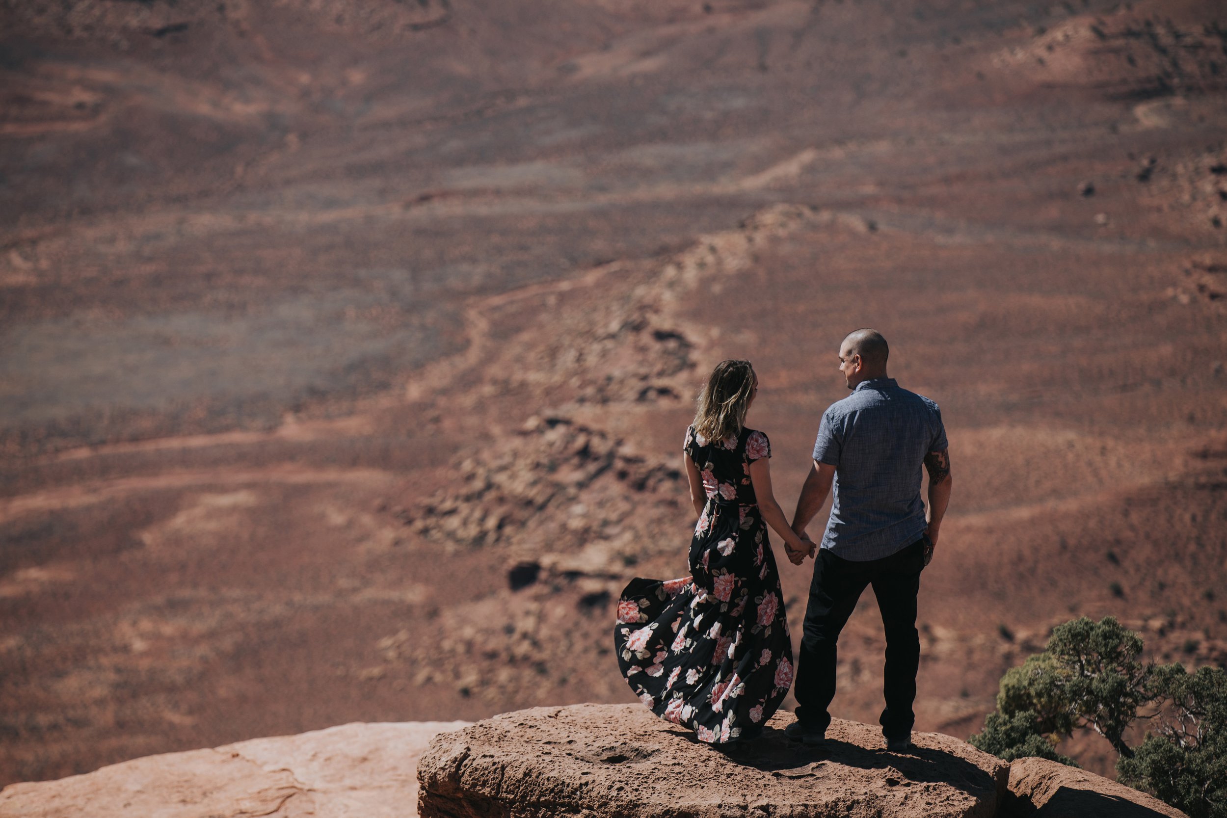 A couple stands on the edge of a canyon, holding hands and gazing into the vast landscape. The woman’s floral dress billows in the wind, creating a picturesque scene of love and connection with nature at sunset.