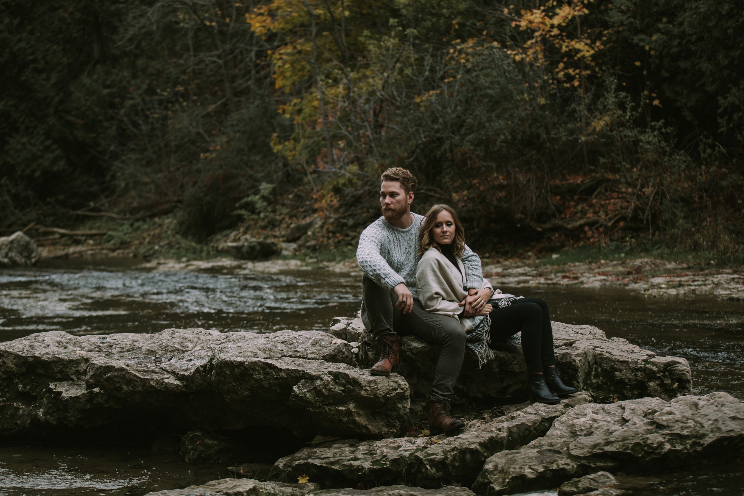 A couple sitting closely together on rocks by a river, surrounded by the warm colours of fall foliage.