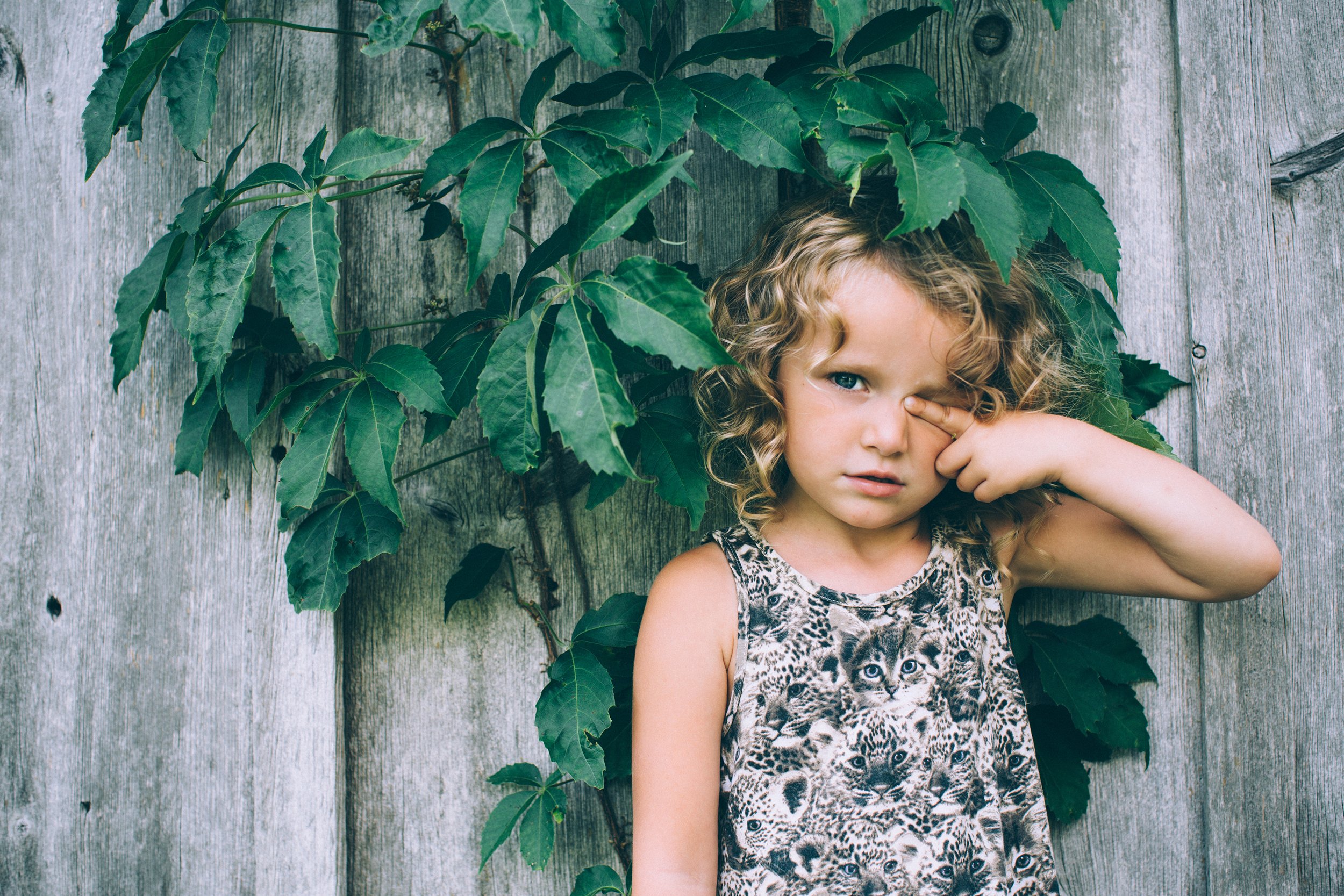 A portrait of a girl with curly hair standing against a rustic wood wall, surrounded by greenery, creating a blend of nature and simplicity.