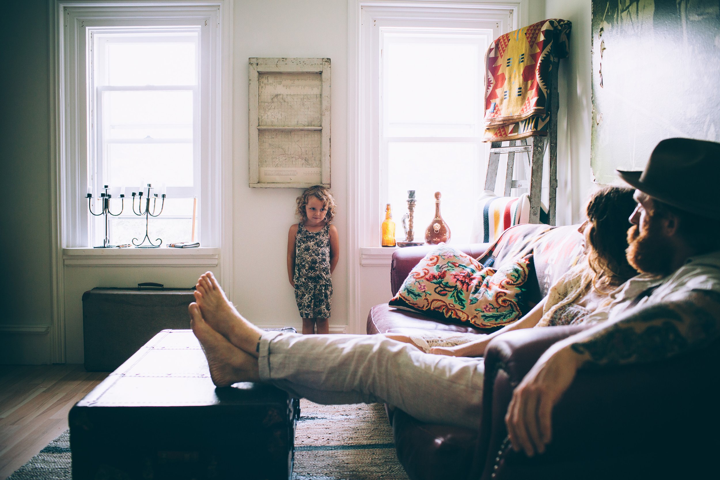 A little girl standing against a wall in a vintage-style living room, with her parents sitting on a couch nearby, creating a nostalgic atmosphere.