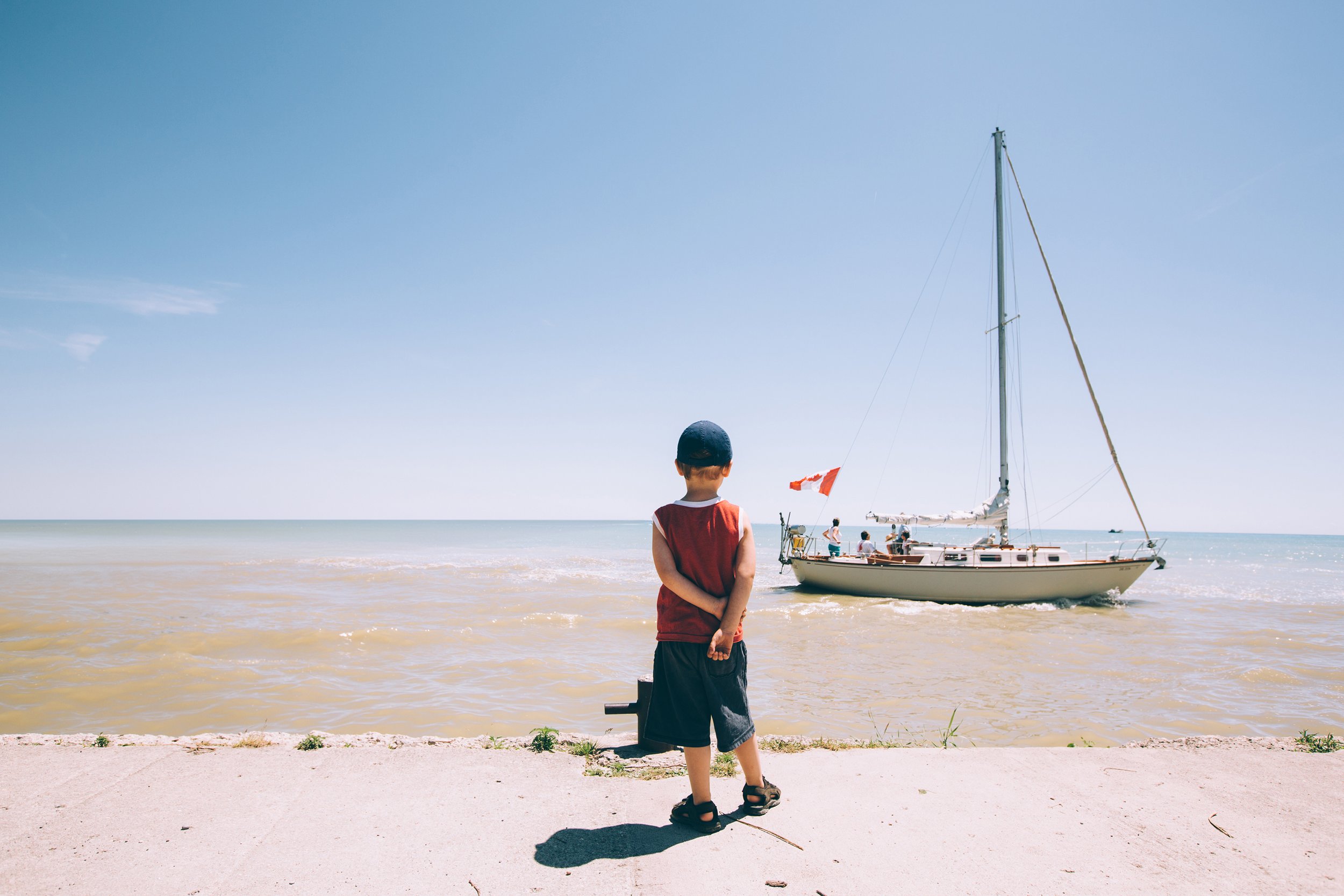 A young boy standing near a harbour on a sunny day, watching a sailboat in the distance.