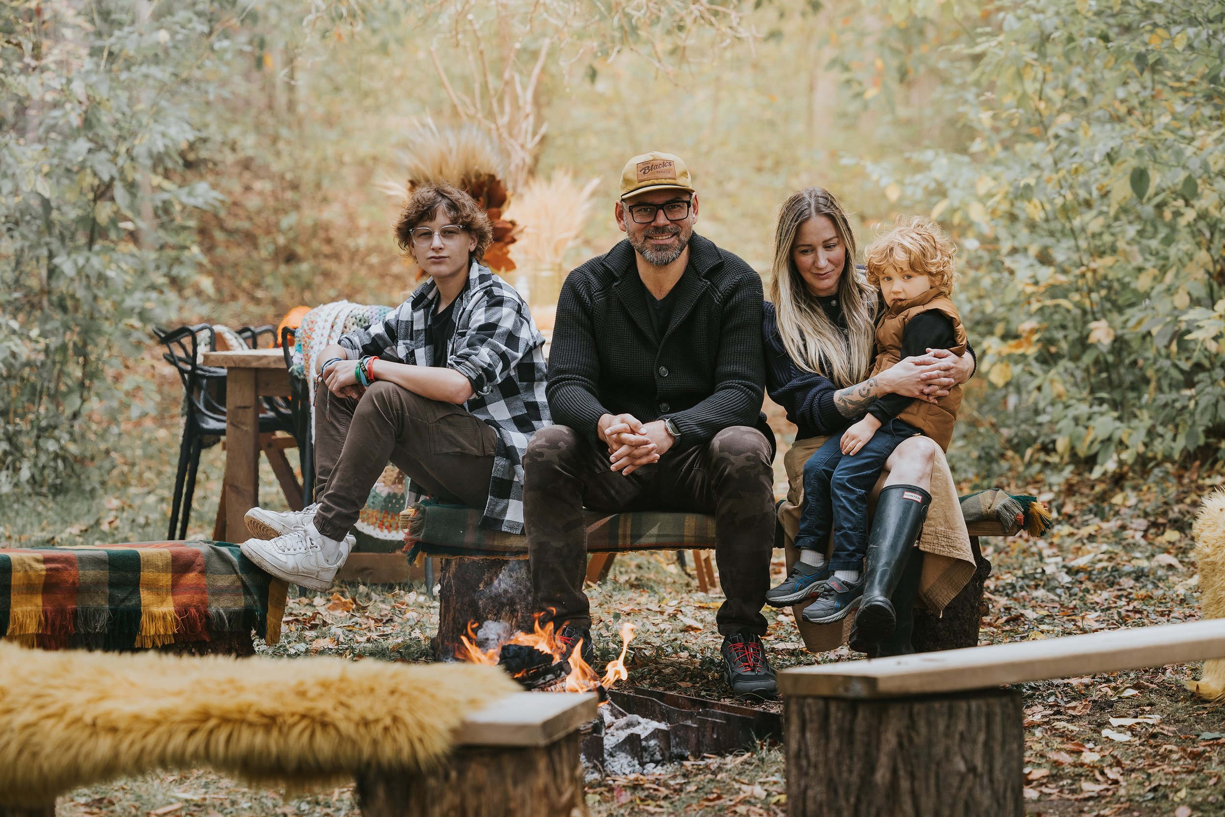 A cozy outdoor family portrait during autumn, with parents and children sitting by a fire pit in the woods.