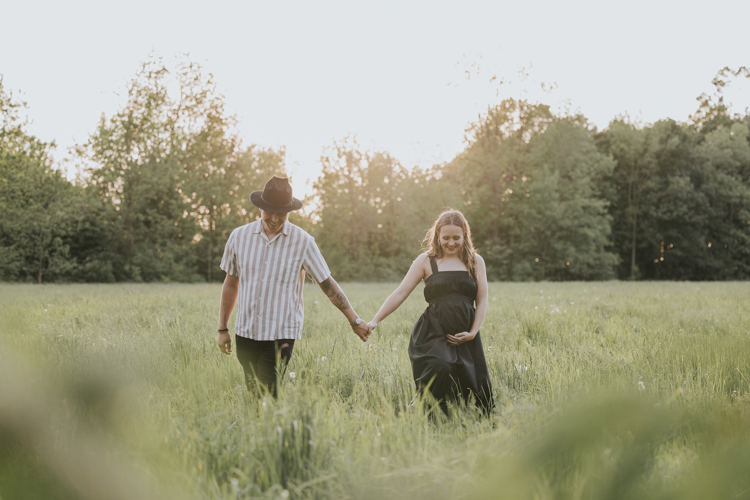 A serene sunset scene of a couple walking hand in hand through a field, with the wife gently cradling her baby bump.